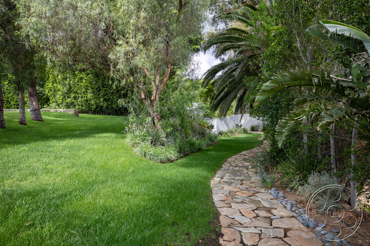 Sinatra House - exterior, natural stone pathway, tropical palm trees, softscape features, landscape garden, drought-resistant plants, lush green grass lawn, meandering garden path, private backyard oasis, irregular stone pavers, curved stone walkway, textured gravel border, landscape shrubs, native plantings, sunlight-dappled area, wooden garden fence, seasonally green foliage