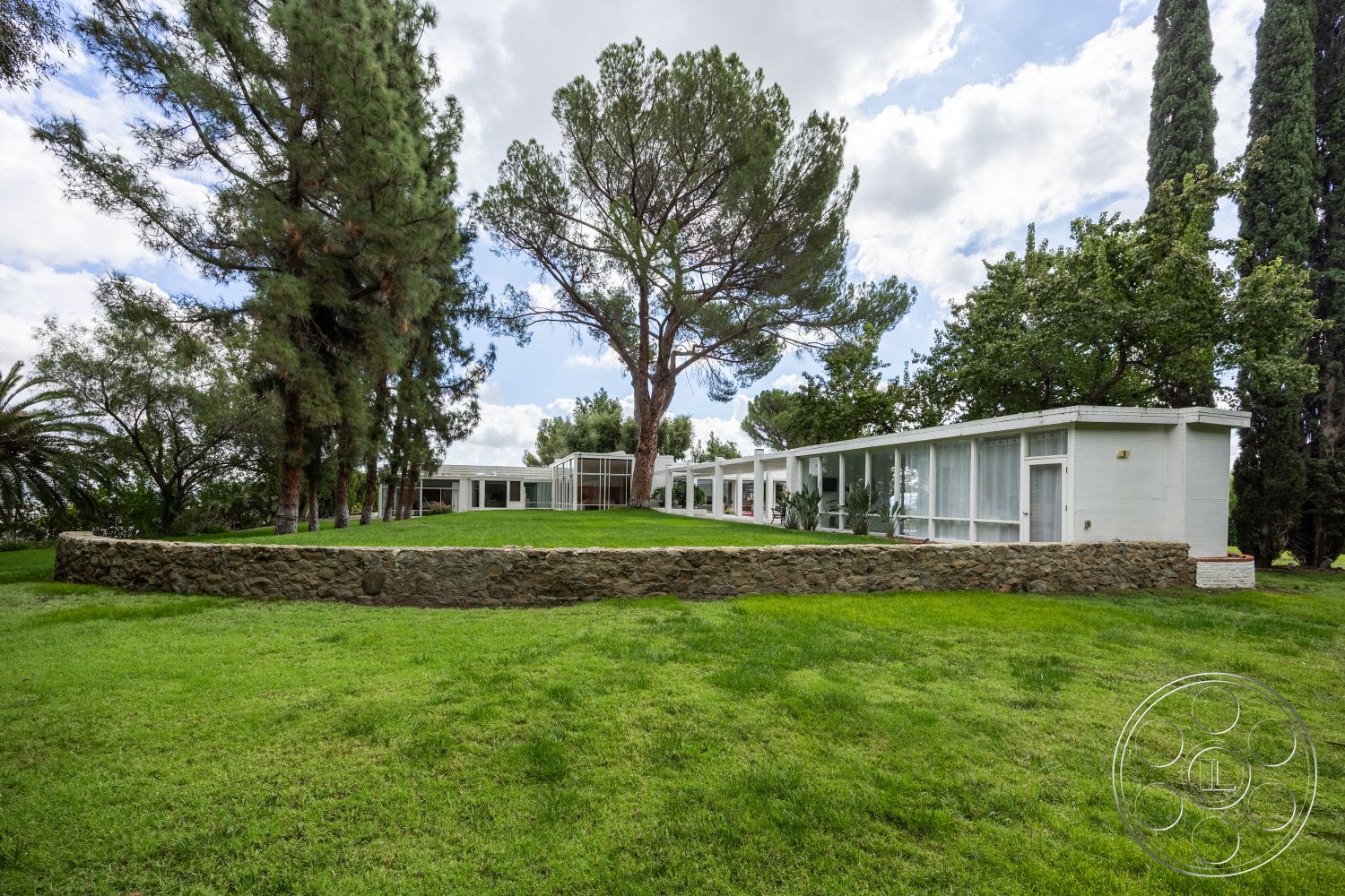 Sinatra House - exterior, low-maintenance landscaping, minimalist design, surrounding greenery, contemporary architecture, landscaped lawn, simple geometric shapes, tall pine trees, stone garden wall, clean lines and angles, natural stone landscaping elements, integrated natural surroundings, flat roof modern home, glass facade design, row of large windows, suburban outdoor setting, open yard space, outdoor space with grass, light-colored wooden cladding, single-story layout