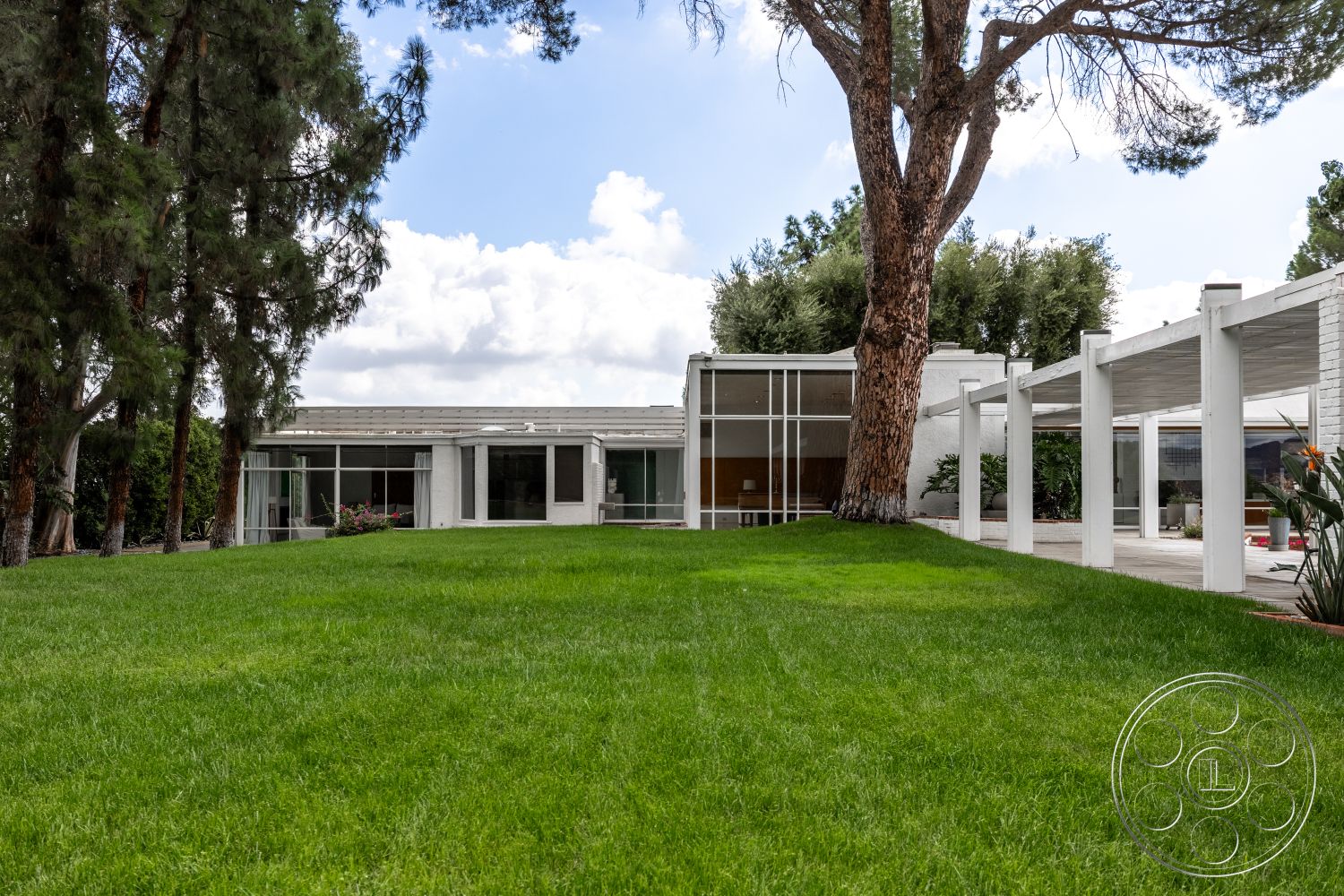 Sinatra House - white stucco walls, open outdoor space, large floor-to-ceiling windows, flat roof design, exposed wood ceiling beams, landscaped garden area, concrete patio flooring, minimalist architectural style, mid-century modern home exterior, grassy lawn setting, patio with pergola structure, surrounding evergreen trees, urban backyard landscape