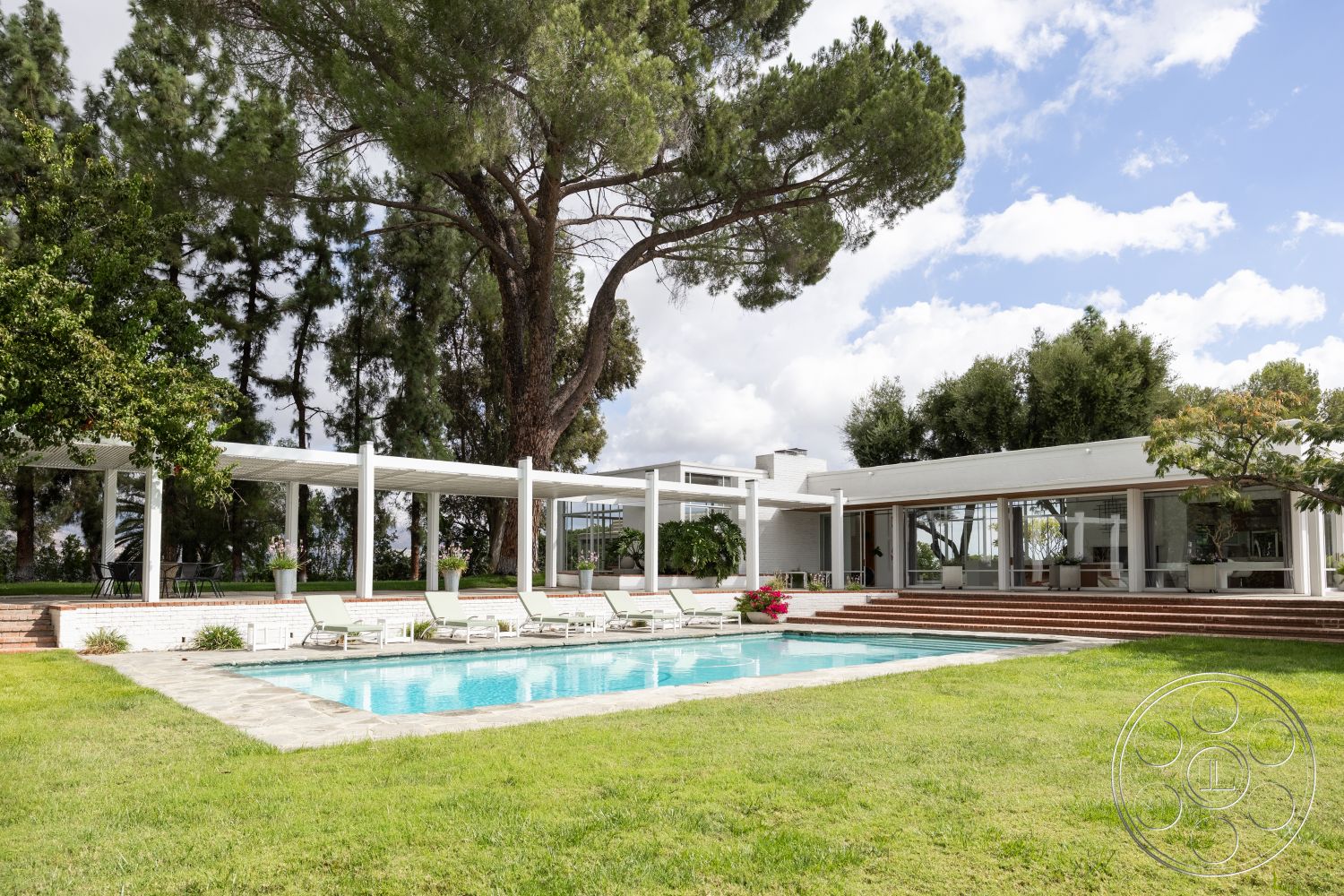 Sinatra House - exterior, white stucco walls, large glass windows, rectangular swimming pool, stone pathway, contemporary outdoor furniture, outdoor lounge area, wooden pergola, minimalist landscaping, sunny outdoor setting, open-air layout, flat roof design, natural grass lawn, modern single-story home, mountain view backdrop, surrounding pine trees, concrete pool surround, brick patio steps