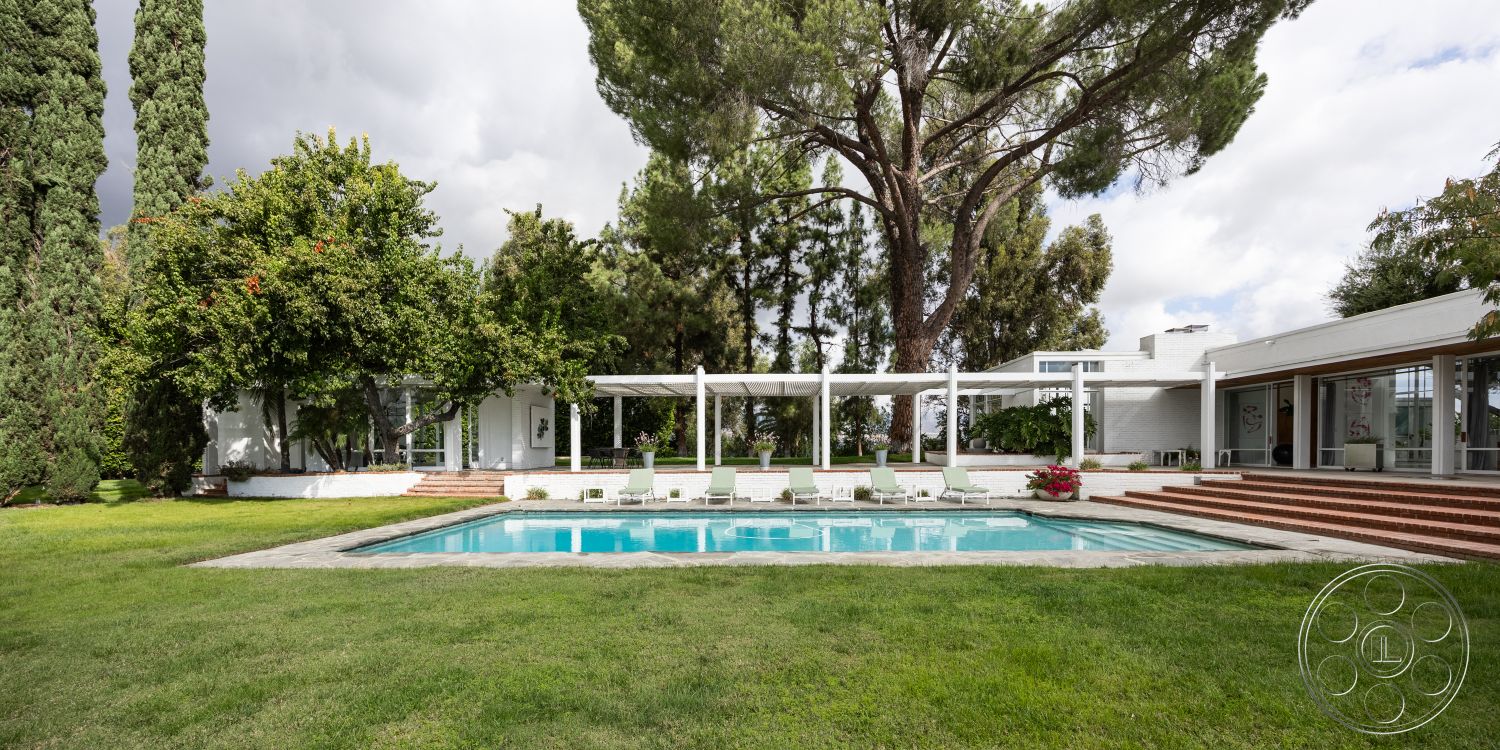 Sinatra House - floor-to-ceiling windows, circular driveway, natural stone pathway, rectangular pool, modern outdoor seating, sunny backyard, exposed wood ceiling beams, landscaped garden area, lush greenery setting, white painted walls, concrete pool border, concrete patio floor, designer outdoor lighting, urban residential exterior, marble pool deck, hardwood decking steps