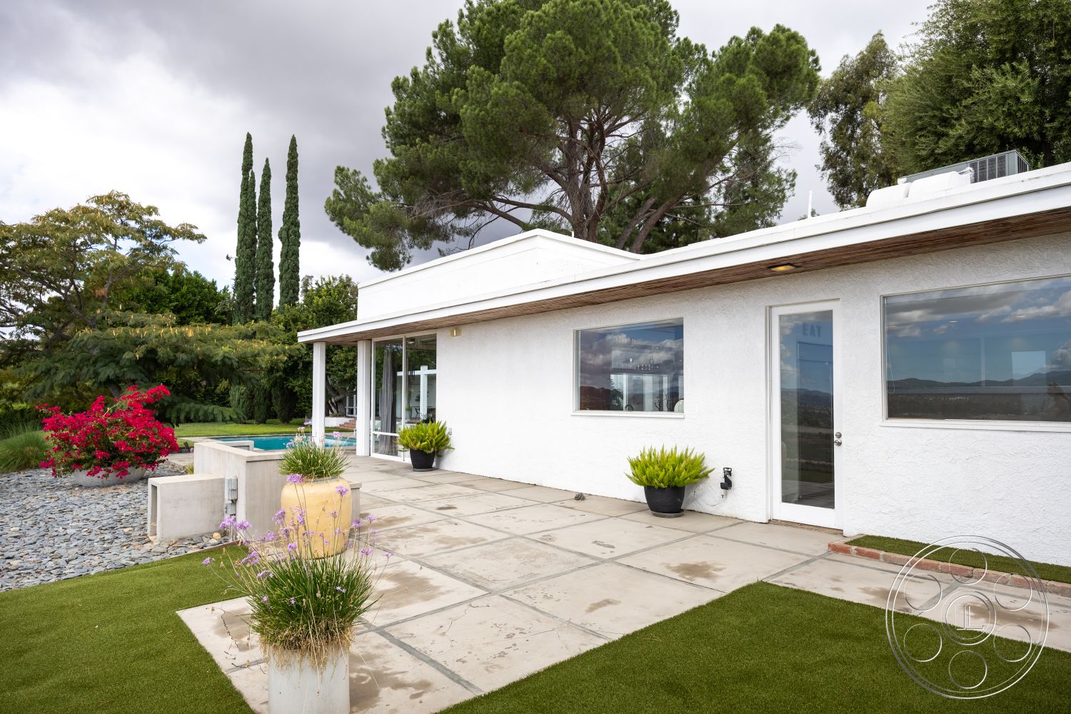 Sinatra House - exterior concrete patio floor, interior open floor plan, exterior white stucco walls, exterior flat roof design, exterior large glass windows, exterior landscaped garden with decorative shrubs, exterior modern L-shaped house, exterior decorative stone border, exterior concrete retaining wall, exterior pool area with rectangular pool, setting suburban backyard, setting mountain view, setting manicured lawn with artificial turf, setting tropical floral accents, interior modern minimalist aesthetic