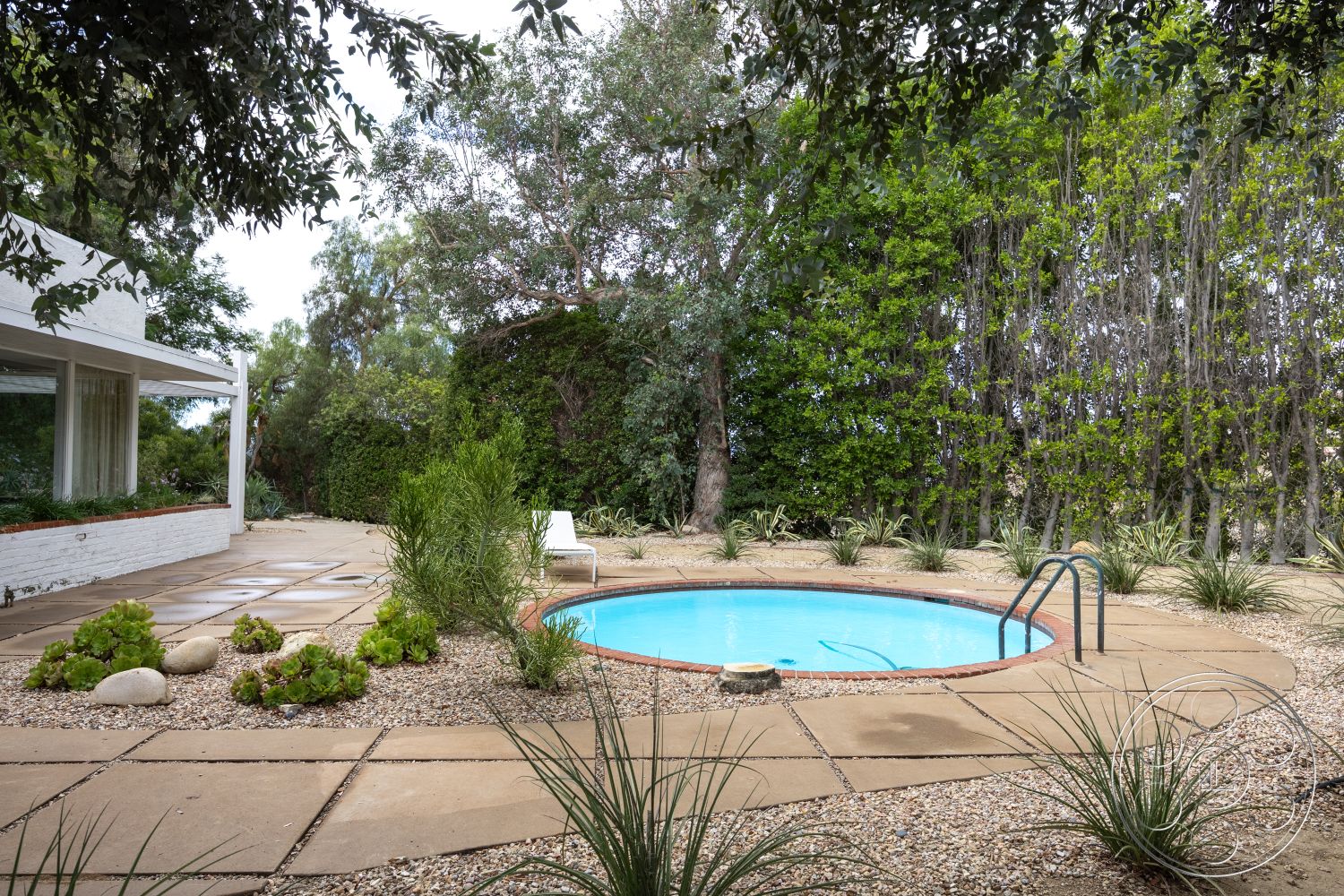 Sinatra House - natural stone accents, potted plants, surrounding greenery, outdoor living space, textured concrete patio, tropical landscaping, modern outdoor seating, circular pool, stone patio flooring, concrete pool border, exterior pool area, shaded outdoor area, poolside relaxation area, decorative gravel ground cover, flagstone pool deck, landscaped garden with succulents, white stucco exterior wall, low maintenance garden design