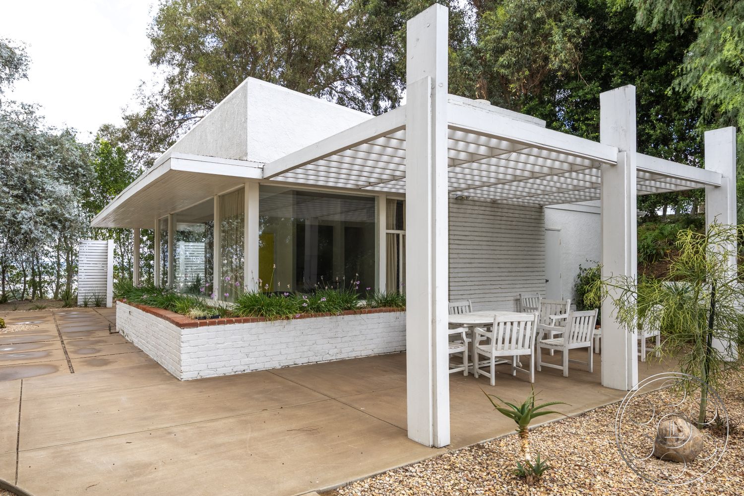 Sinatra House - open concept layout, large floor-to-ceiling windows, smooth concrete driveway, flat roof design, white painted exterior walls, urban garden setting, minimalist architectural style, exposed concrete patio flooring, red brick planter border, pergola outdoor seating area, modern rectangular table and chairs, surrounded by landscaped greenery, exterior with clean lines, extensions for natural light, sculptural landscape elements