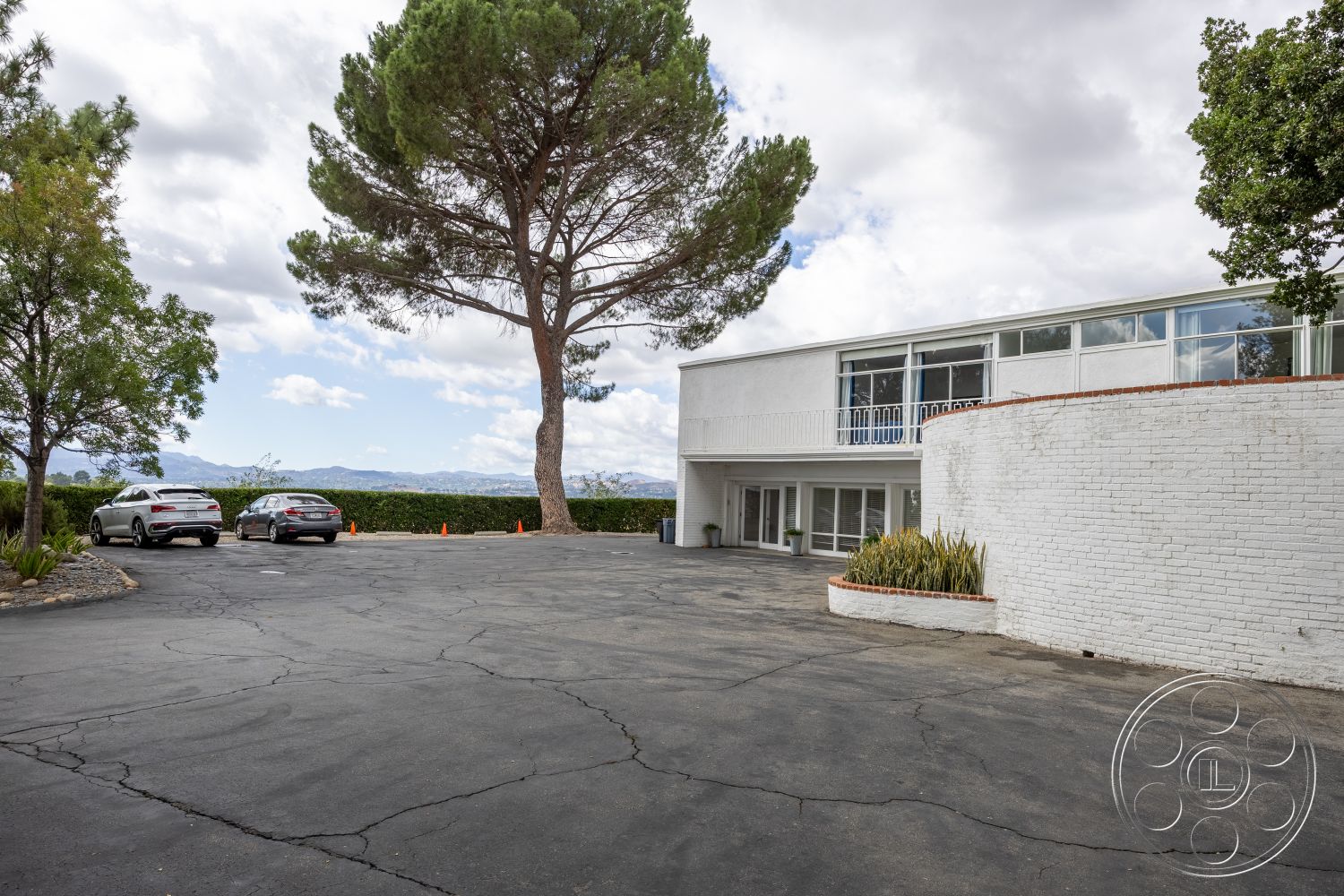 Sinatra House - large glass windows, urban landscape, modern architectural style, curved driveway, asphalt driveway, outdoor parking area, flat roof design, landscaped garden area, decorative stone border, minimalist facade, mountain view backdrop, white brick exterior walls, exposed concrete foundation, aluminum balcony railing, tree-lined outdoor setting, exterior entryway patio, interior living spaces, horizontal window alignment