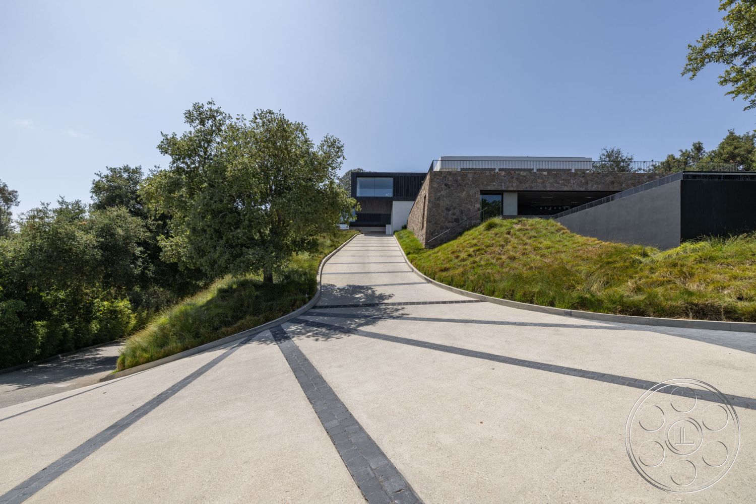 Modern 513 - minimalist architectural style, exterior driveway with concrete surface, curved driveway with stone borders, modern hillside home with grass landscaping, exposed stone wall on architecture, large glass windows for natural lighting, steel railing details on steps, section of grass for sustainable landscaping, vaulted design elements in modern construction, urban hillside setting with views, landscape integration with native vegetation