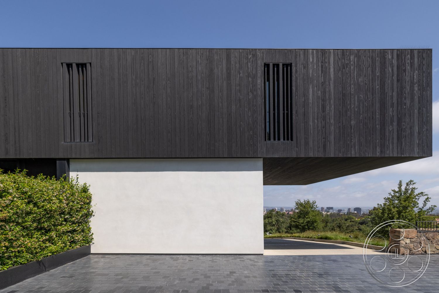 Modern 513 - exterior, slate stone driveway, contemporary structure, flat roof architecture, wooden facade cladding, smooth white concrete walls, black wood beam overhang, horizontal slatted wooden window coverings, geometric modern design, landscaped greenery in foreground, urban skyline background