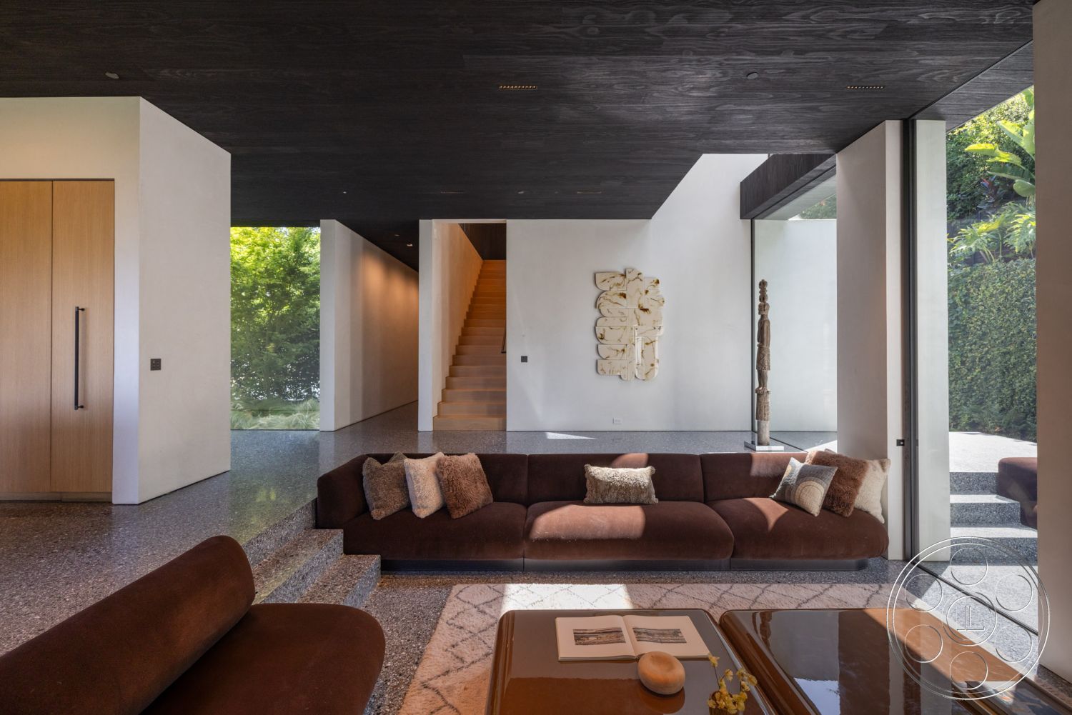 Modern 513 - interior neutral color palette, interior stone feature wall, interior concrete floor with terrazzo finish, interior exposed wood ceiling, interior large floor-to-ceiling glass windows, interior contemporary brown sectional sofa, interior custom wood cabinetry, interior textured wall art installation, interior minimalist staircase with wood treads, interior natural light-filled open layout, interior polished concrete landing, interior soft textile throw pillows, interior decorative coffee table, interior integration of nature with surrounding greenery