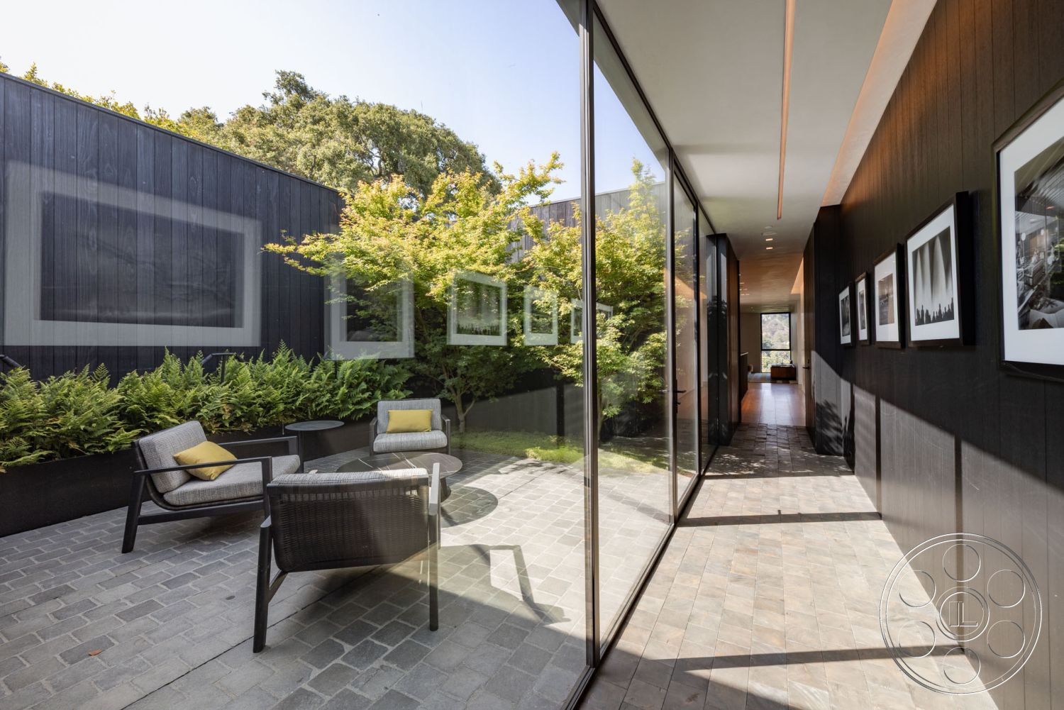 Modern 513 - sustainable building materials, minimalist architectural style, urban residential environment, exterior patio with stone paver flooring, modern glass walls with floor-to-ceiling windows, wood paneling on exterior walls, contemporary low-maintenance landscaped garden, open-air courtyard design with greenery, modern outdoor seating area with metal-framed chairs, natural light-filled hallway leading to garden, artistic wall with multiple framed photographs, textured concrete walkway adjacent to garden, smooth concrete planter with greenery, Zen-inspired garden setting, scenic views of surrounding trees