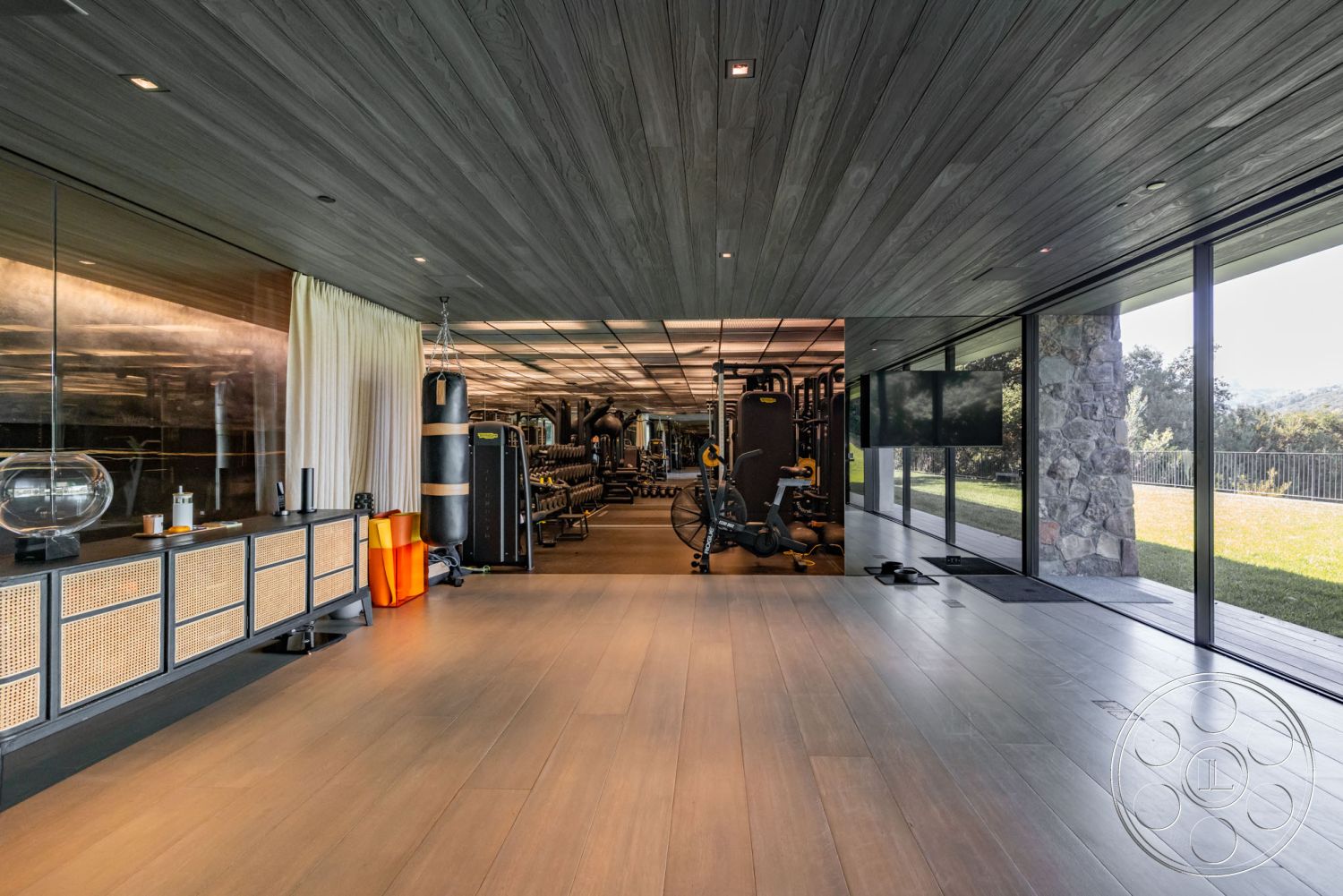 Modern 513 - minimalist design elements, seamless indoor-outdoor connection, recessed ceiling lighting, neutral color palette with warm tones, exposed wood ceiling planks, interior hardwood gym flooring, glass wall partition with views, contemporary home gym space, integrated storage cabinets with cane detail, modern training equipment area, natural light-filled fitness room, stone feature wall accent, open exercise area with rolling grass views, sliding glass doors to garden patio, luxurious home workout environment