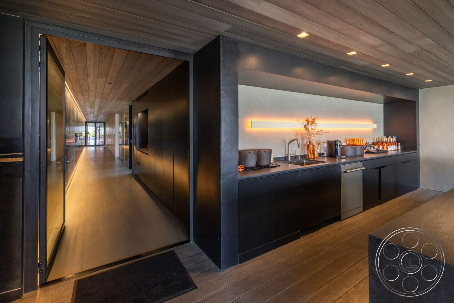 Modern 513 - modern minimalist aesthetic, built-in kitchen appliances, black metal door hardware, concrete kitchen countertops, neutral color palette with warm accents, spacious hallway with natural light, interior kitchen with black cabinetry, textured plaster wall behind kitchen, floating shelves with kitchenware, warm wood ceiling panels, glass entry door with metal frame, direct lighting along the hallway, hardwood flooring in kitchen and hallway, open concept design with views to outdoor, contemporary dining area visible from kitchen, floor-to-ceiling windows in hallway, LED strip lighting above countertops, stylish beverage bar setup, seamless transition from kitchen to hallway