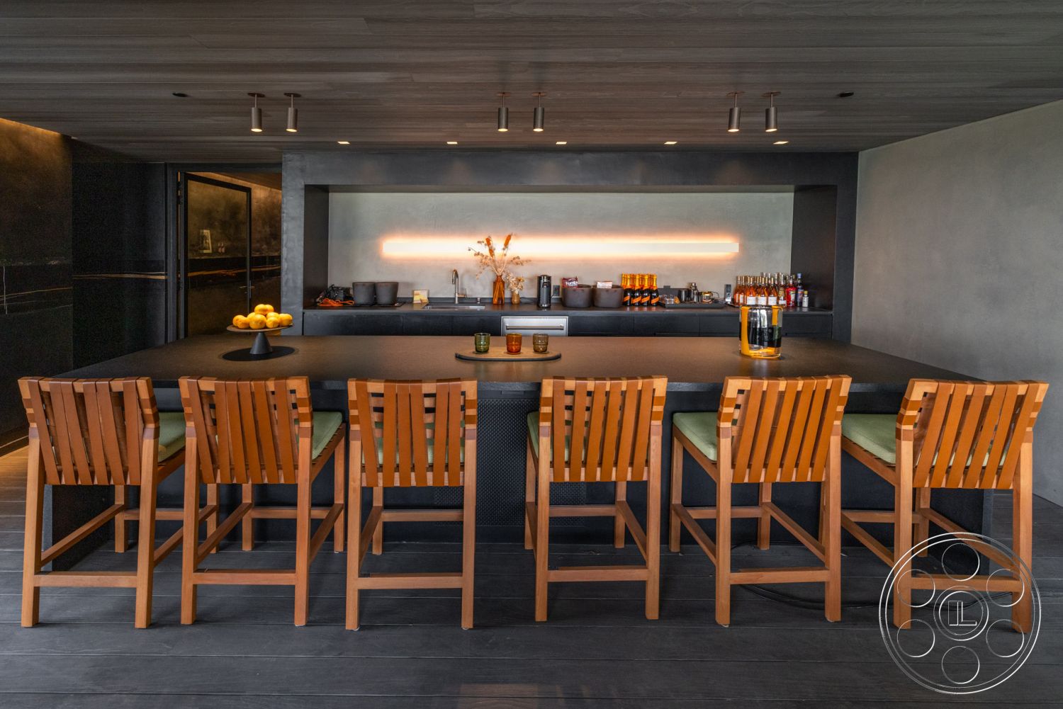 Modern 513 - exposed wood ceiling panels, minimalist design with clean lines, interior kitchen countertop with black stone surface, interior bar area with wooden bar stools, modern lighting fixtures above bar, concrete wall treatment behind bar, open floor plan kitchen and living area, dark cabinetry with seamless finish, green cushion seating on bar stools, decorative vase with dried flowers on countertop, floor-to-ceiling glass doors for natural lighting, neutral color palette with contrasting elements, textured plaster wall beside bar, subtle decorative elements on bar shelves, high-end kitchen appliances integrated into cabinetry, ambient LED lighting behind bar area