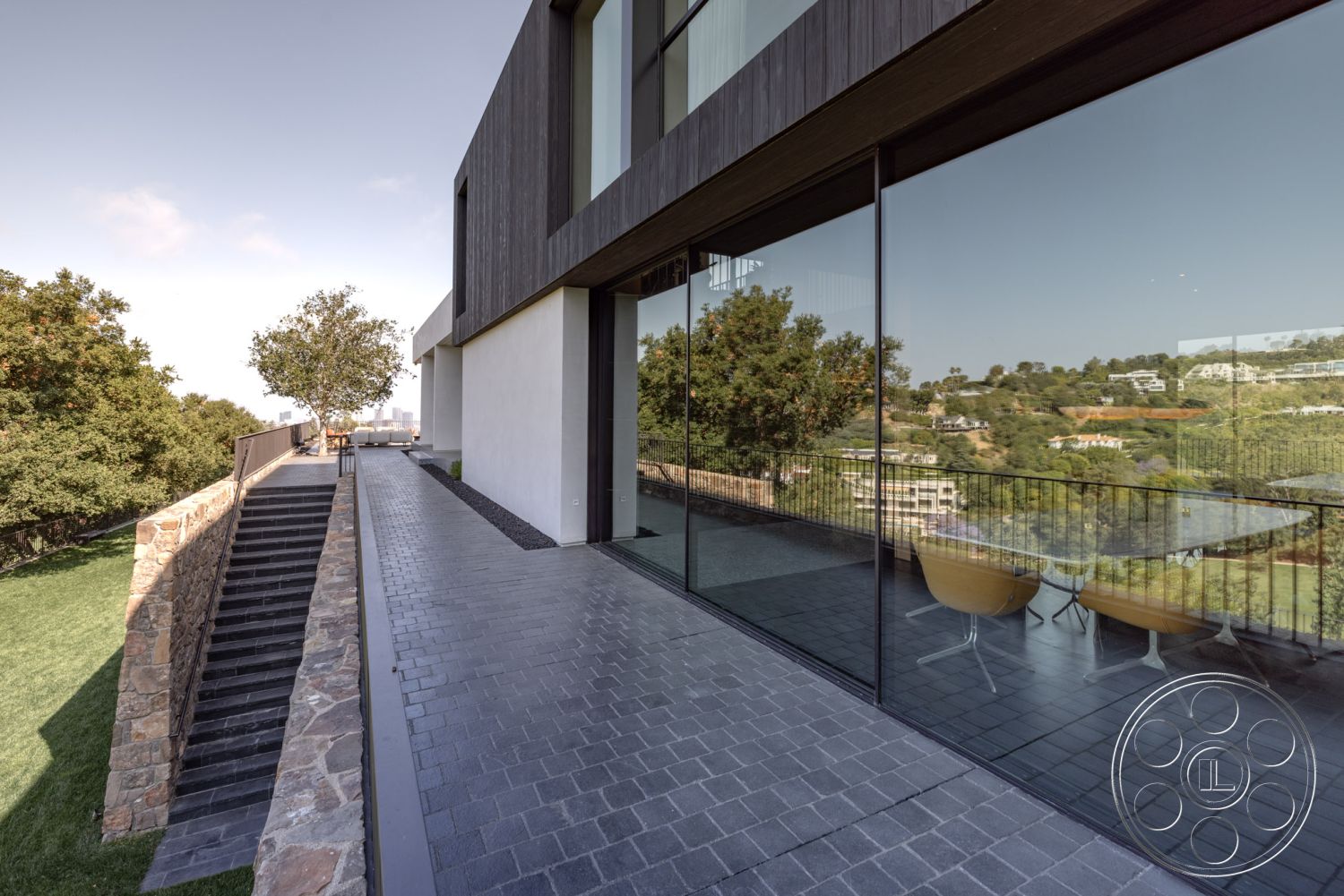 Modern 513 - exterior, large glass sliding doors, city skyline view, stone retaining wall, natural stone textures, horizontal wood siding, landscaped garden area, concrete patio flooring, modern minimalist design, open outdoor dining area, staggered stone steps, rectangular balcony with railings, planted green lawn, geometric exterior shapes, flat roof profile