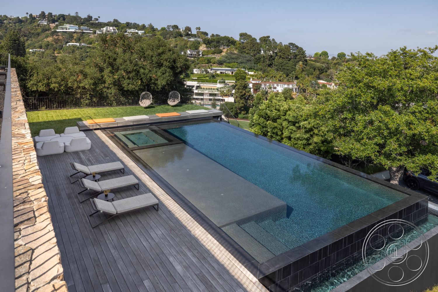 Modern 513 - exterior infinity pool with glass edge, hardwood deck surrounding pool area, modern lounge chairs with cushions, concrete pool coping and border, landscaped hillside setting with trees, city view from elevated location, circular stone paver patio, built-in hot tub within pool area, open-air relaxation space, rope swing chairs for outdoor seating