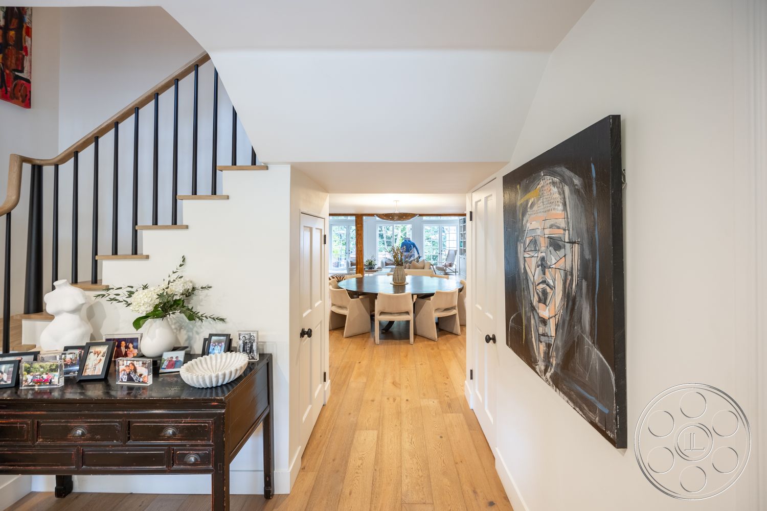 Americana 201 - round dining table, contemporary dining area, recessed lighting fixtures, interior hallway, natural light-filled space, decorative vase with flowers, wood beam ceiling, white painted walls, hardwood entryway floors, modern artwork on wall, black metal stair railings, exposed wood staircase, glass dining room windows, gallery-style photo display, open-concept interior design