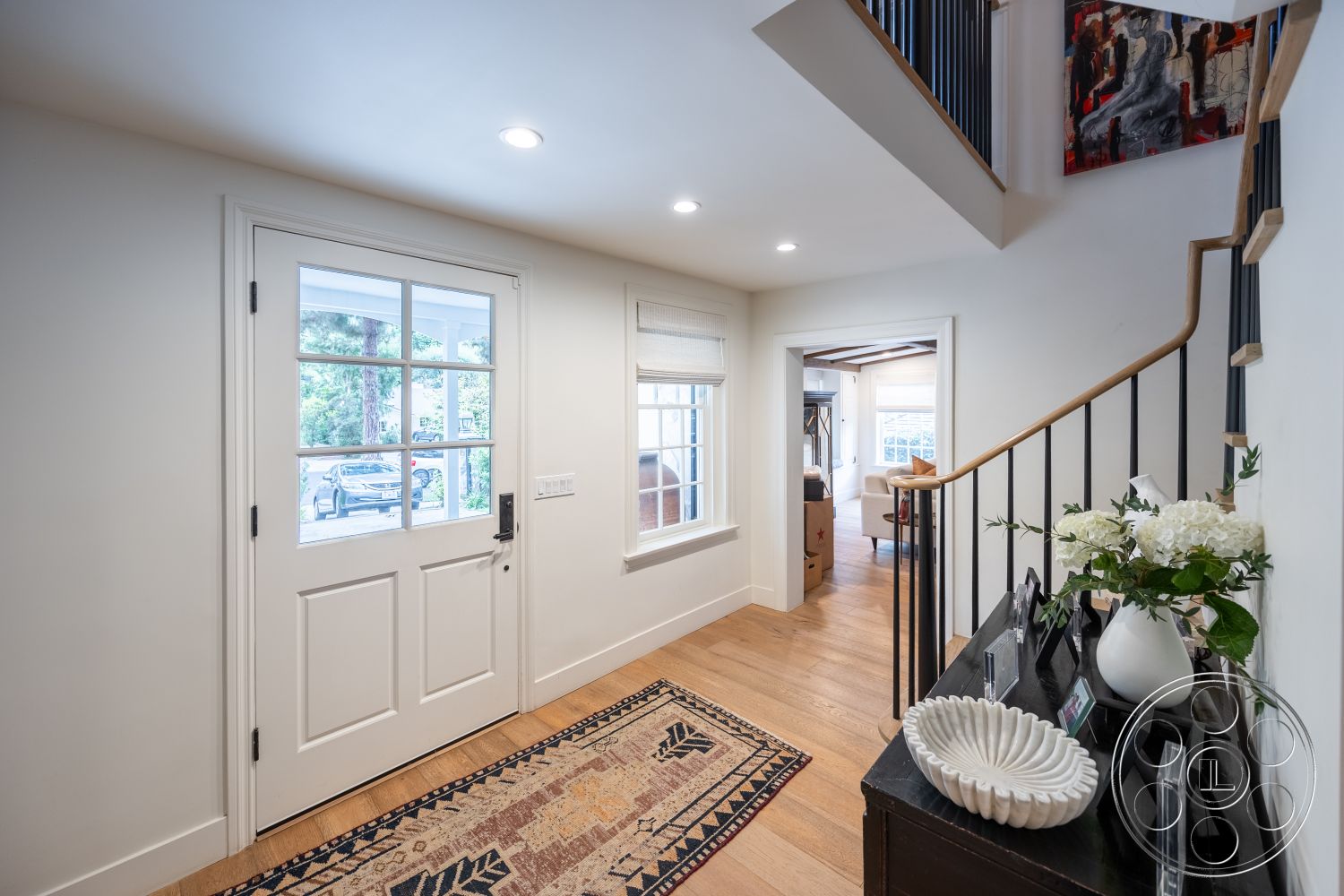 Americana 201 - natural light through large windows, white painted walls, interior entrance hallway, black metal stair railings, hardwood flooring in entryway, modern wooden front door with glass panes, soft beige area rug with intricate patterns, decorative console table, vase with white hydrangeas, open concept design leading to living area, artwork on wall above stairway, cozy seating area visible through doorway