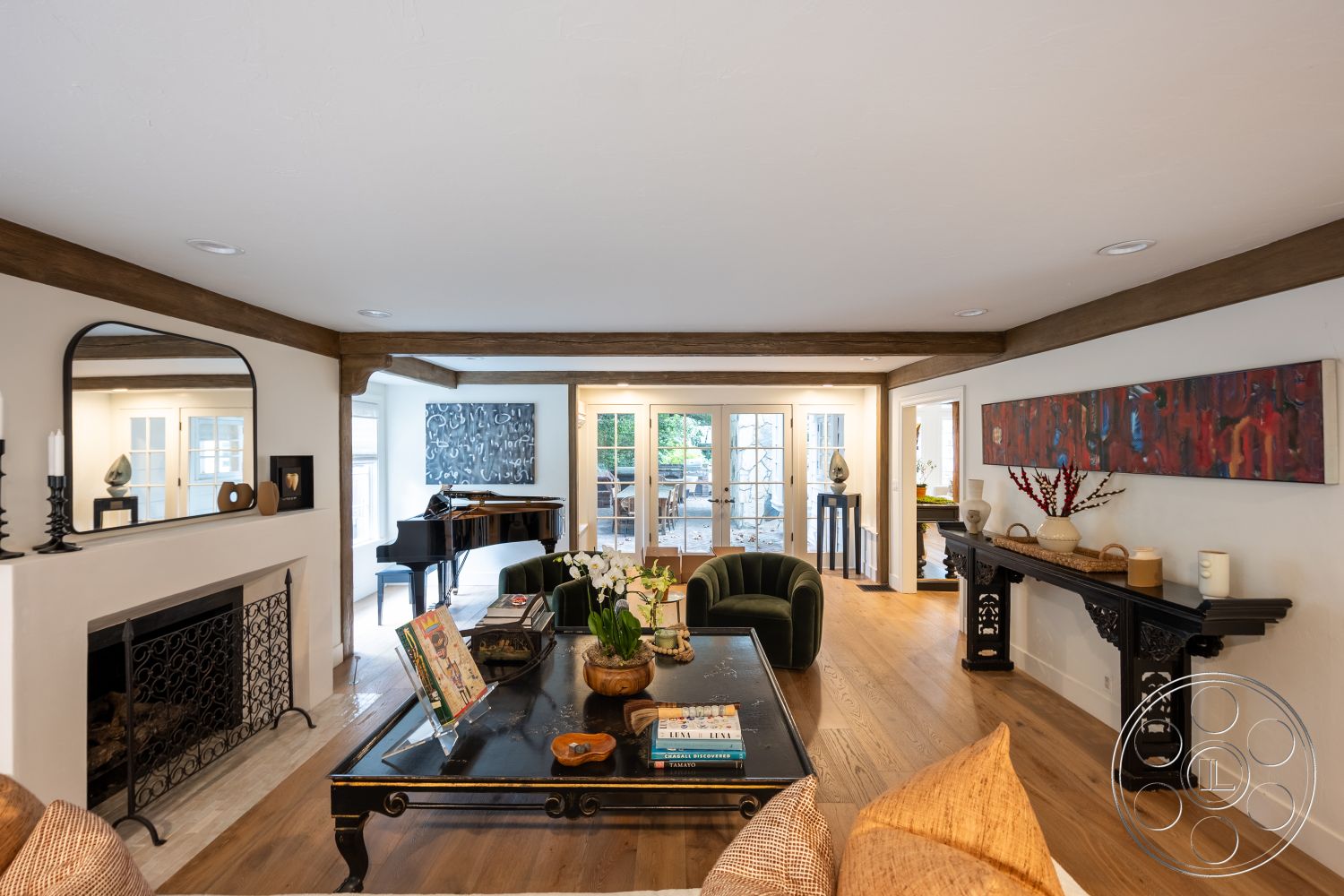 Americana 201 - round coffee table, black piano, textured throw pillows, comfortable seating area, exposed wood ceiling beams, hardwood living room floors, white painted walls, open floor plan design, stone fireplace surround, modern chandelier lighting, vintage furniture accents, large mirror accent, traditional fireplace grate, glass door access to patio, art piece wall decoration, floral centerpiece decor, suburban indoor setting, natural light flood, contemporary art style, black lacquered console table