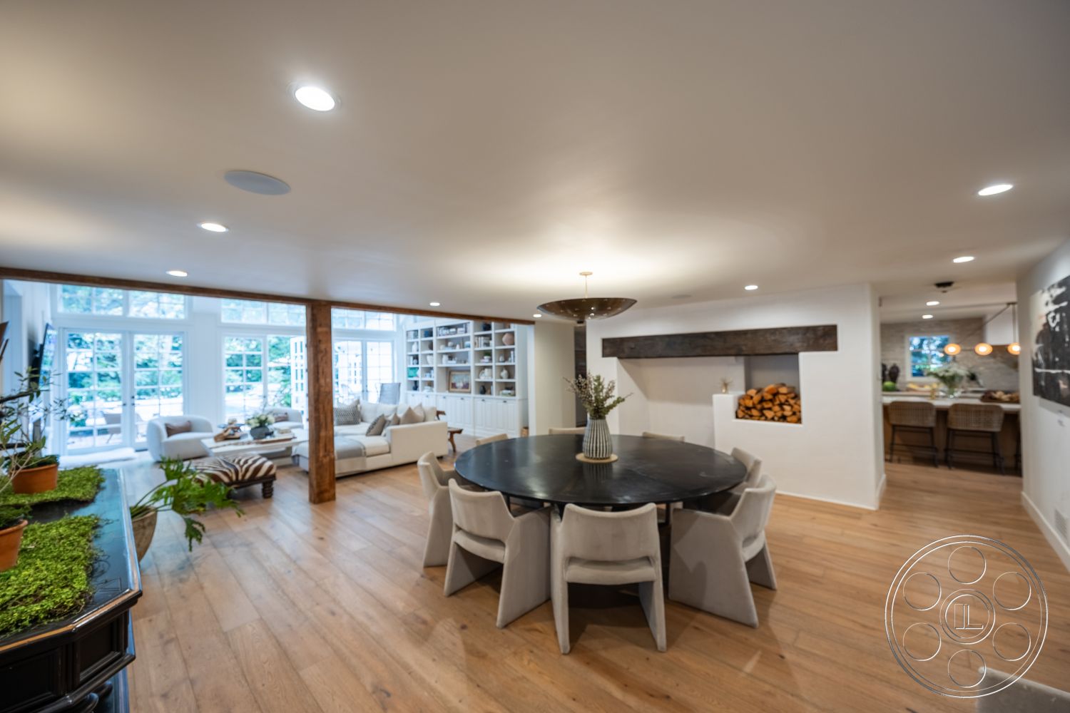 Americana 201 - neutral color palette, round black dining table, exposed wood ceiling beams, hardwood living room floors, cozy dining nook, suburban backyard setting, open floor plan layout, large windows with garden view, stylish pendant lighting, contemporary sofa with throw pillows, natural light-filled interior, interior living area, textured white plaster walls, stone fireplace feature wall, modern upholstered dining chairs, built-in bookcase with decor, rustic wood log storage, indoor plants for added greenery