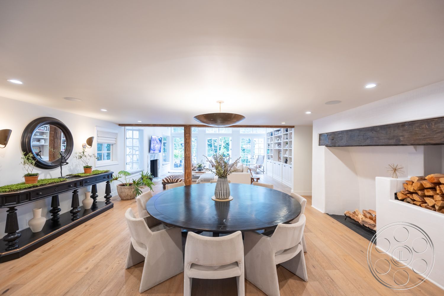 Americana 201 - neutral color palette, large windows, natural light, hardwood flooring, cozy atmosphere, firewood storage, plant decor, open floor plan, decorative plants, exposed wood beams, round dining table, white plaster walls, contemporary lighting fixture, built-in bookshelves, modern dining chairs, black wooden sideboard