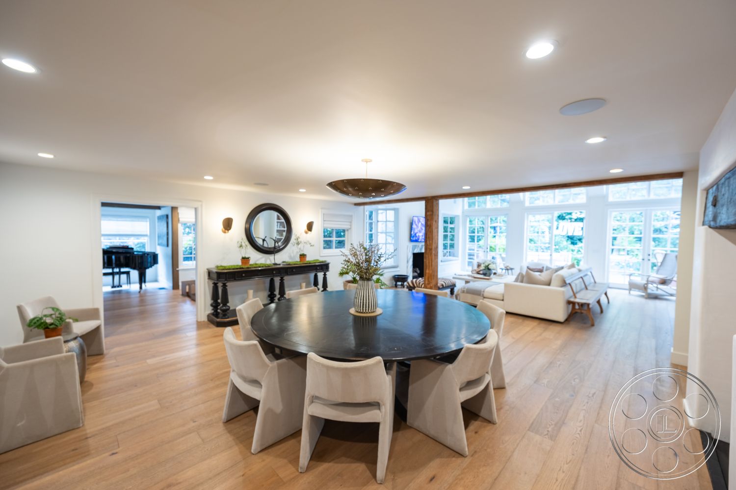 Americana 201 - wall-mounted television, modern pendant lighting, natural light-filled space, white painted walls, hardwood flooring in living area, exposed wooden beam ceiling, round dining table with upholstered chairs, black dining table finish, large windows with garden views, contemporary decorative mirror, stylish black sideboard, open-concept living room, cozy sofa seating area