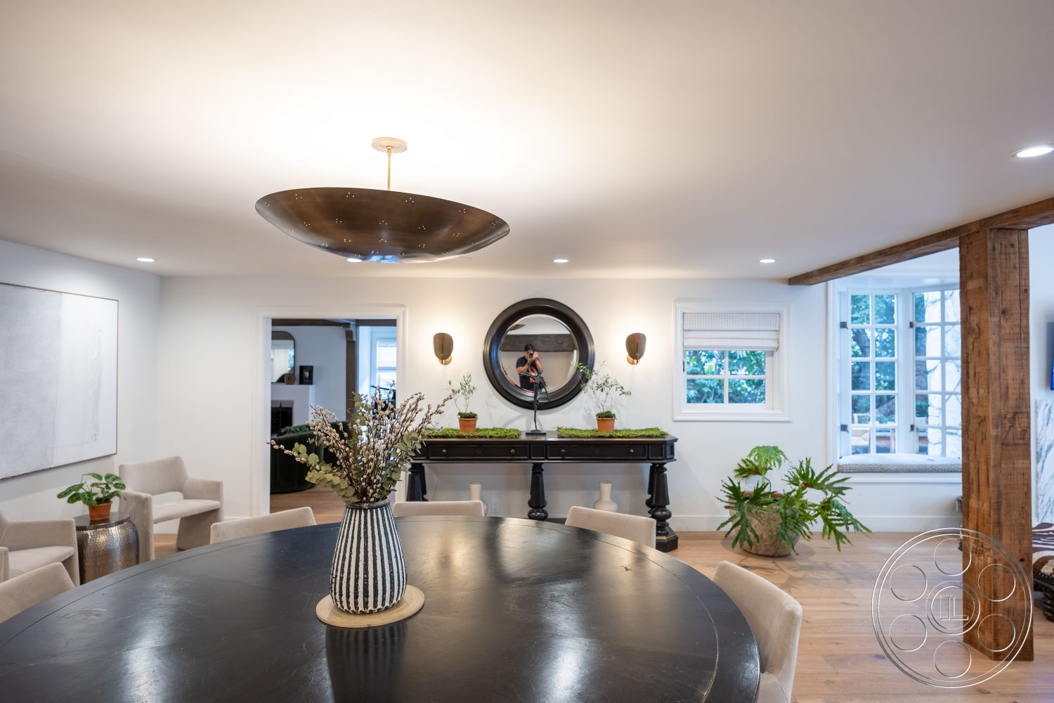 Americana 201 - neutral color palette, modern pendant lighting, upholstered dining chairs, exposed wood beams, open concept living space, black round dining table, interior dining room, white painted walls, large windows with natural light, framed artwork on wall, hardwood flooring in living area, round wall mirror, decorative black console table, potted plant décor, plant features in room