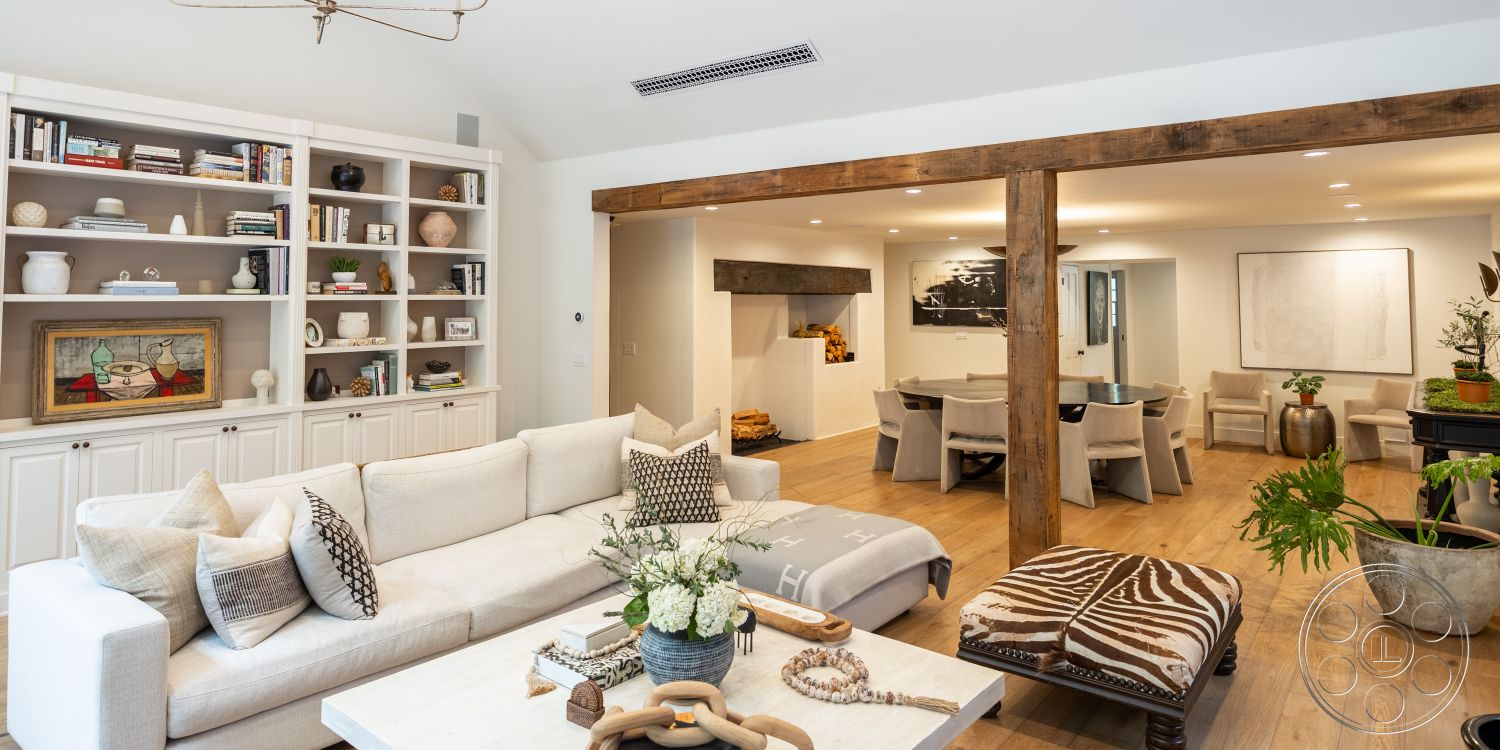 Americana 201 - interior hardwood living room floors, interior exposed wood ceiling beams, interior muted color palette, interior cozy atmosphere, interior white painted walls, interior decorative throw pillows, interior natural light from large windows, interior circular coffee table, interior built-in shelves, interior soft sectional sofa, interior rustic wood beam doorway, interior abstract art wall decoration, interior patterned accent chair, interior open concept living and dining area, interior decorative plant stands, interior side table with decorative objects, interior neutral-colored area rug, interior light fixture with vintage design, interior inviting living space, interior modern farmhouse style