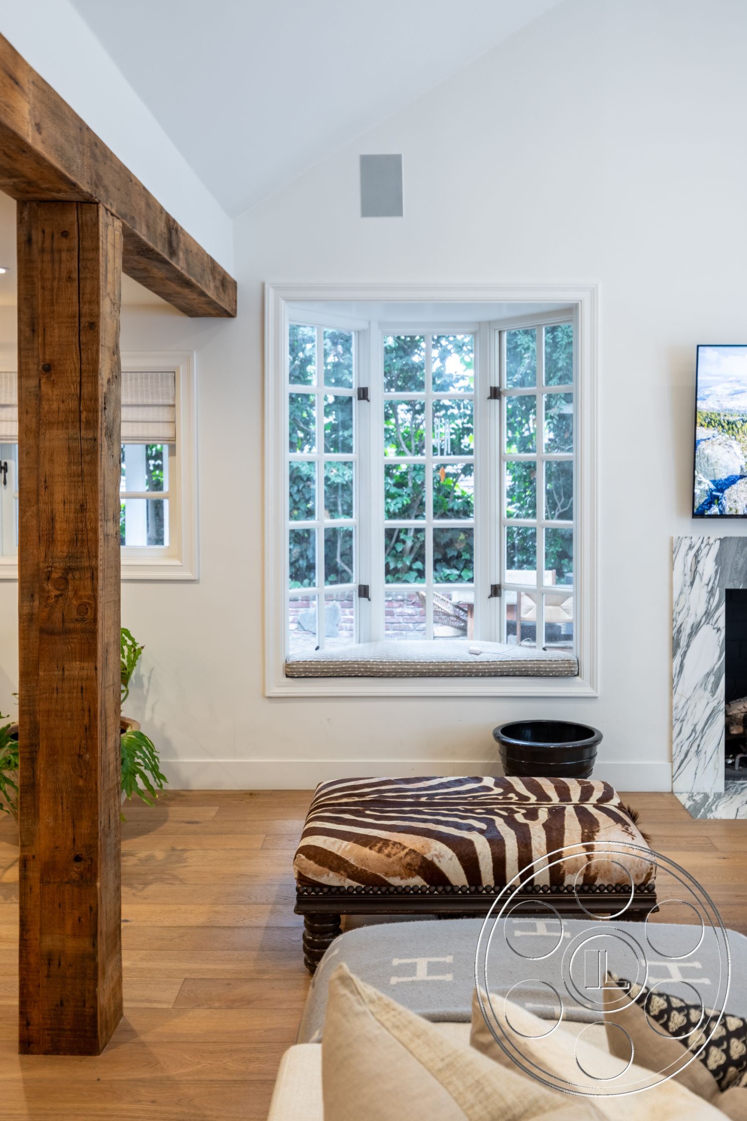 Americana 201 - inviting atmosphere, open floor plan, functional layout, decorative throw pillows, contemporary interior design, stylish home decor, natural light interior, white painted walls, marble fireplace surround, cozy living area, rustic wood accents, exterior window with wooden trim, hardwood living room floor, exposed wood beam column, textured fabric ottoman, bay window seat with cushions