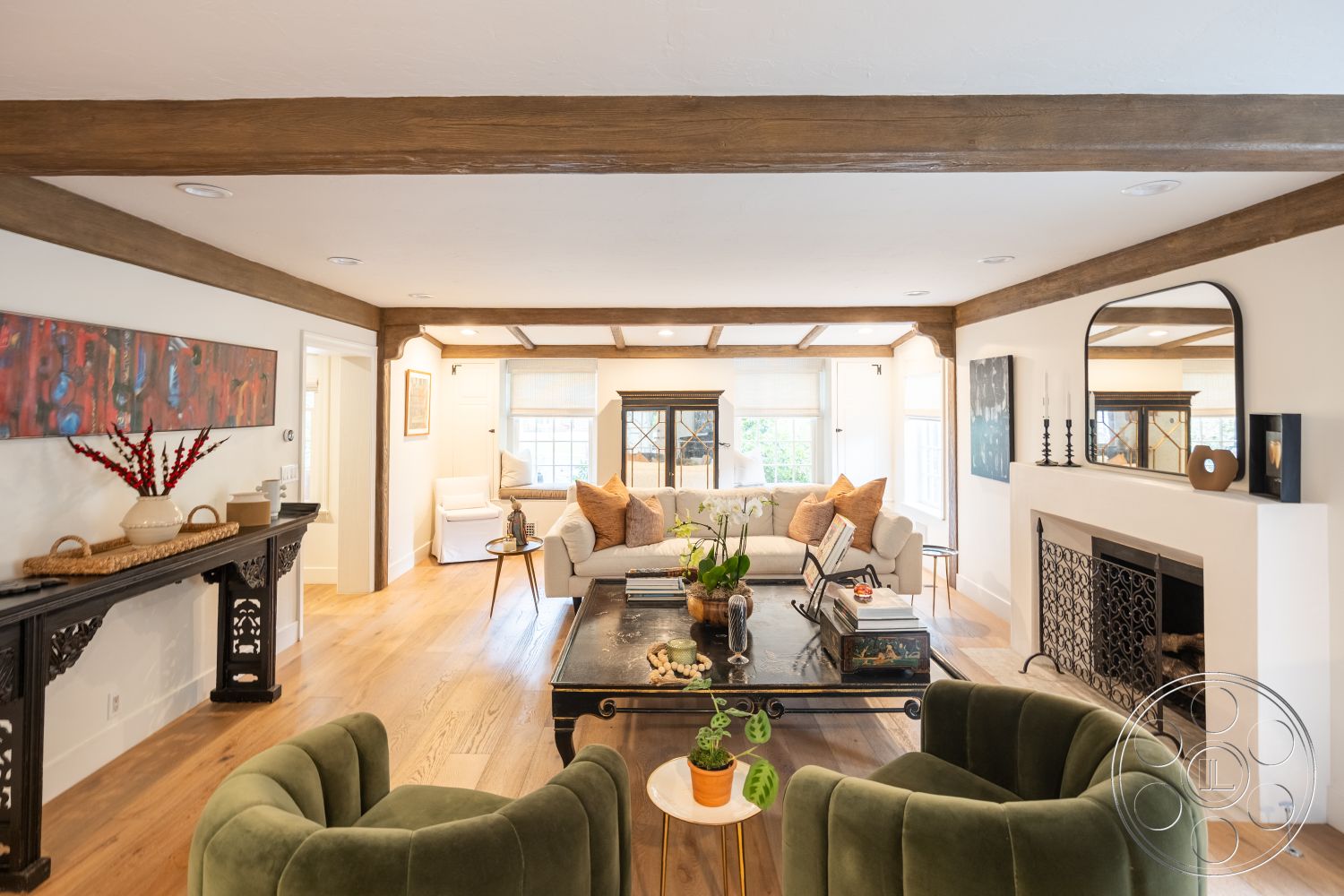 Americana 201 - neutral color palette, textured wall art, large windows for natural light, modern coffee table, exposed wood ceiling beams, white painted walls, hardwood living room flooring, open living area layout, decorative fireplace with metal screen, traditional console table, upholstered sofa with decorative pillows, green velvet accent chairs