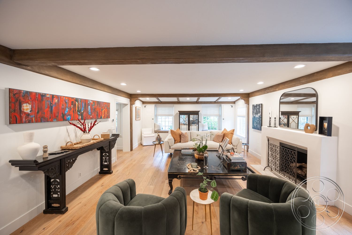 Americana 201 - natural light windows interior, exposed wood ceiling beams interior, white painted walls interior, open floor plan living space interior, light hardwood flooring living room, black accent table interior, round coffee table interior, cushioned armchairs interior, neutral-colored sofa interior, colorful abstract wall art interior, decorative fireplace surround interior, crown molding details interior, glass-top side table interior, textural throw pillows interior