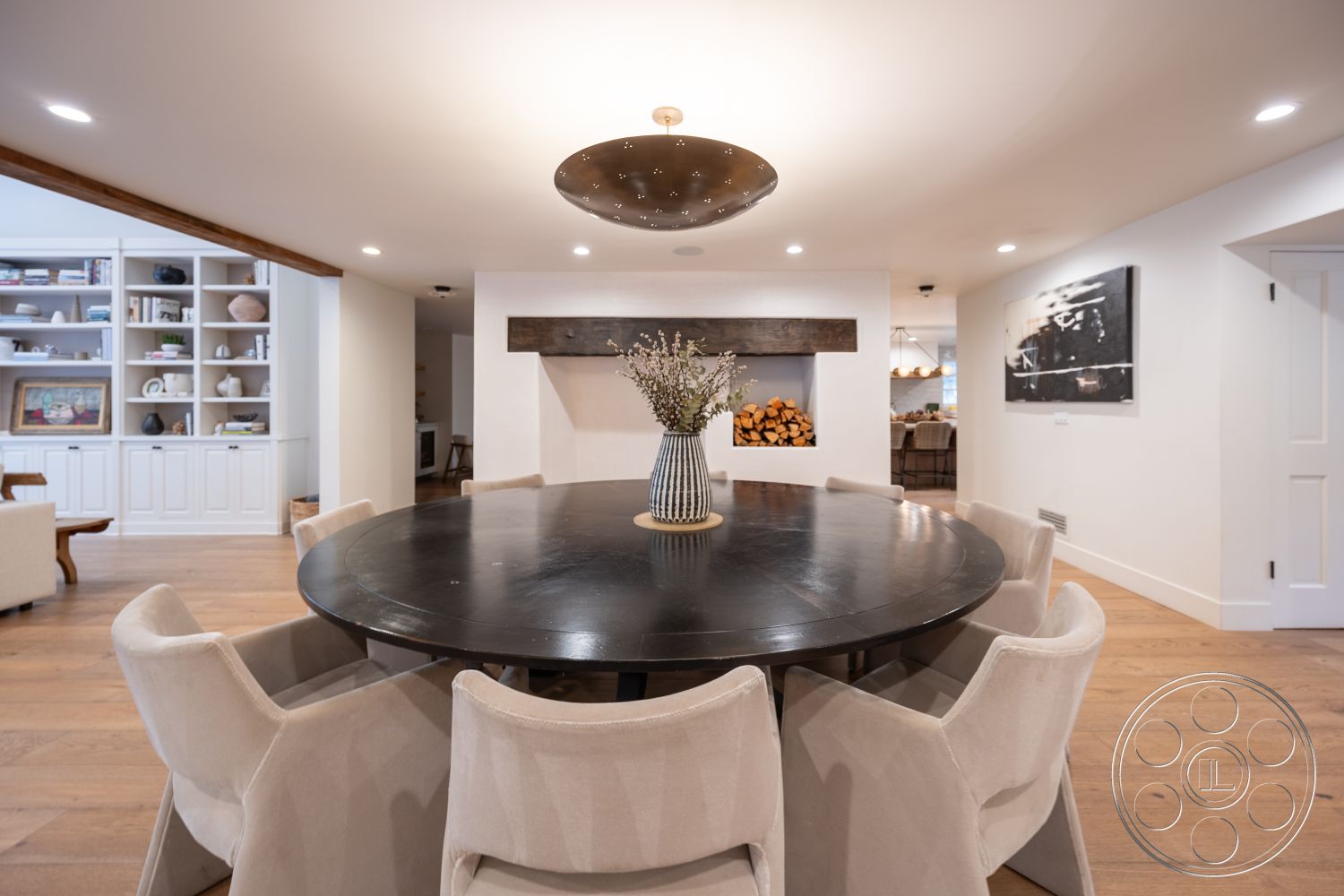 Americana 201 - neutral color palette, textured wall art, open floor plan, modern pendant lighting, upholstered dining chairs, interior dining area, white painted walls, exposed wood beam ceiling, contemporary home design, hardwood dining room flooring, round black wood dining table, built-in cabinetry with shelving, firewood storage niche, cozy indoor setting, natural light from adjacent rooms