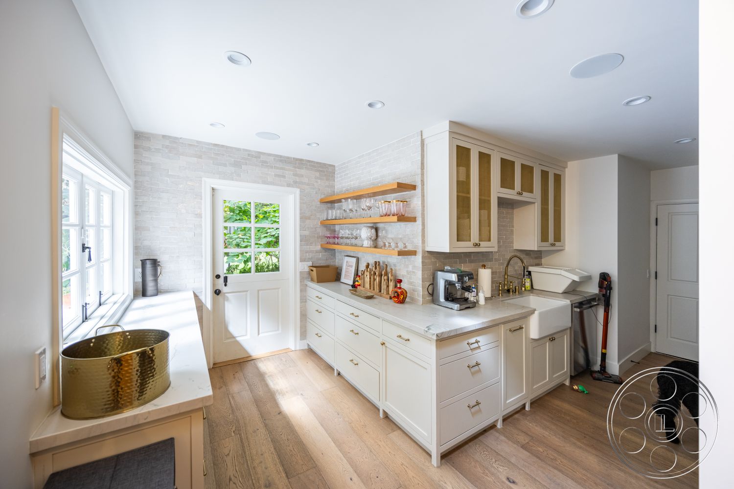 Americana 201 - neutral color palette, modern pendant lighting, open space design, white shiplap walls, interior kitchen, high ceilings with recessed lighting, light wood plank flooring, contemporary kitchen layout, gray stone backsplash, white cabinetry with glass inserts, natural wood floating shelves, stainless steel sink with farmhouse style, subway tile accent wall, traditional white door with window, windows with garden view