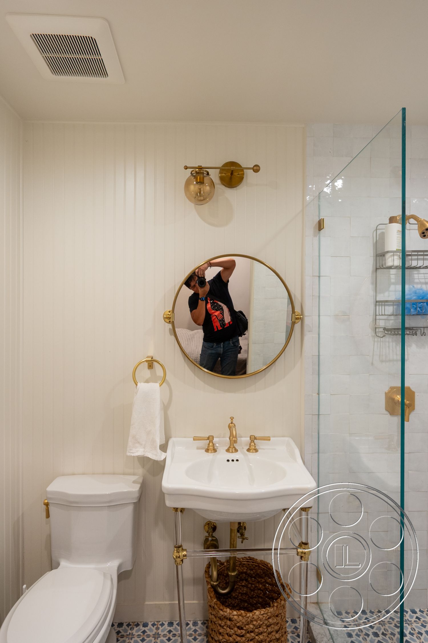 Americana 201 - glass shower enclosure, white porcelain toilet, woven storage basket, modern faucet design, interior bathroom, minimalist bathroom decor, freestanding porcelain sink, brass faucet details, gold round mirror, hexagon marble shower tiles, beadboard wall treatment, gold wall sconces, textured white towels, decorative mosaic floor tiles, plumbing visible under sink