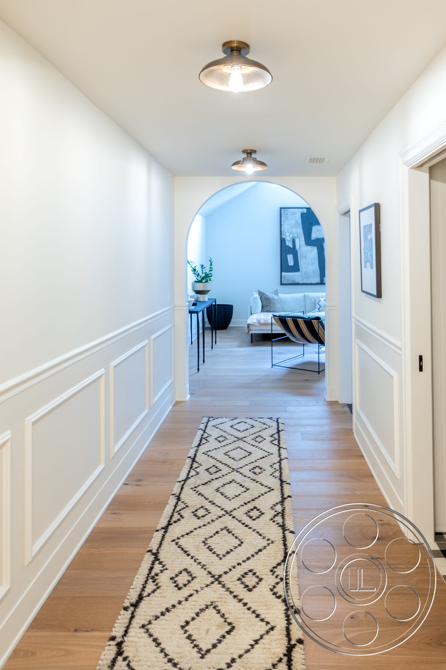 Americana 201 - natural light, minimalist design, light hardwood flooring, modern lighting fixtures, open concept layout, decorative wall art, arched doorway, patterned area rug, interior hallway, white painted walls, indoor plant decoration, wainscoting wall treatment