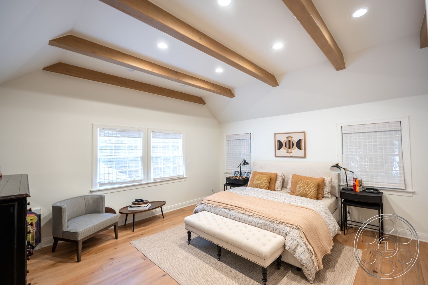 Americana 201 - textured area rug, vaulted ceiling, master bedroom, tufted bench, cozy reading nook, light-colored bedding, glass table lamp, exposed wood ceiling beams, natural light interior, hardwood bedroom floors, white painted walls, artwork wall decor, modern bed with upholstered headboard, picture windows with blinds, cotton throw blanket, minimalist chair, contemporary nightstand