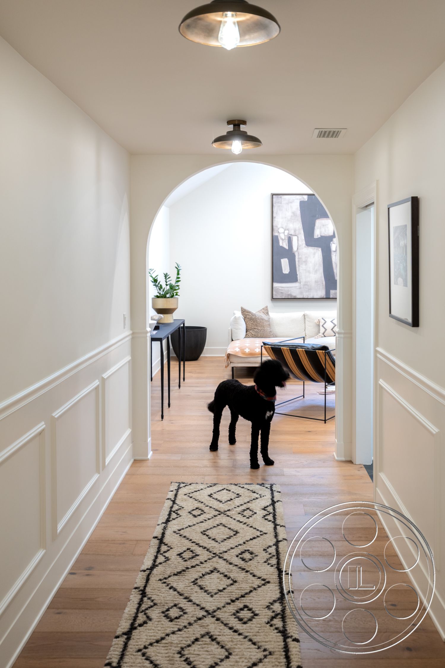 Americana 201 - contemporary art on walls, modern pendant lighting fixtures, minimalist furniture arrangement, interior hallway with hardwood flooring, textured area rug with geometric patterns, white painted walls with wainscoting, arched doorway leading to living space, open floor plan to adjacent living room, decorative potted plant in corner, black dog as a design element