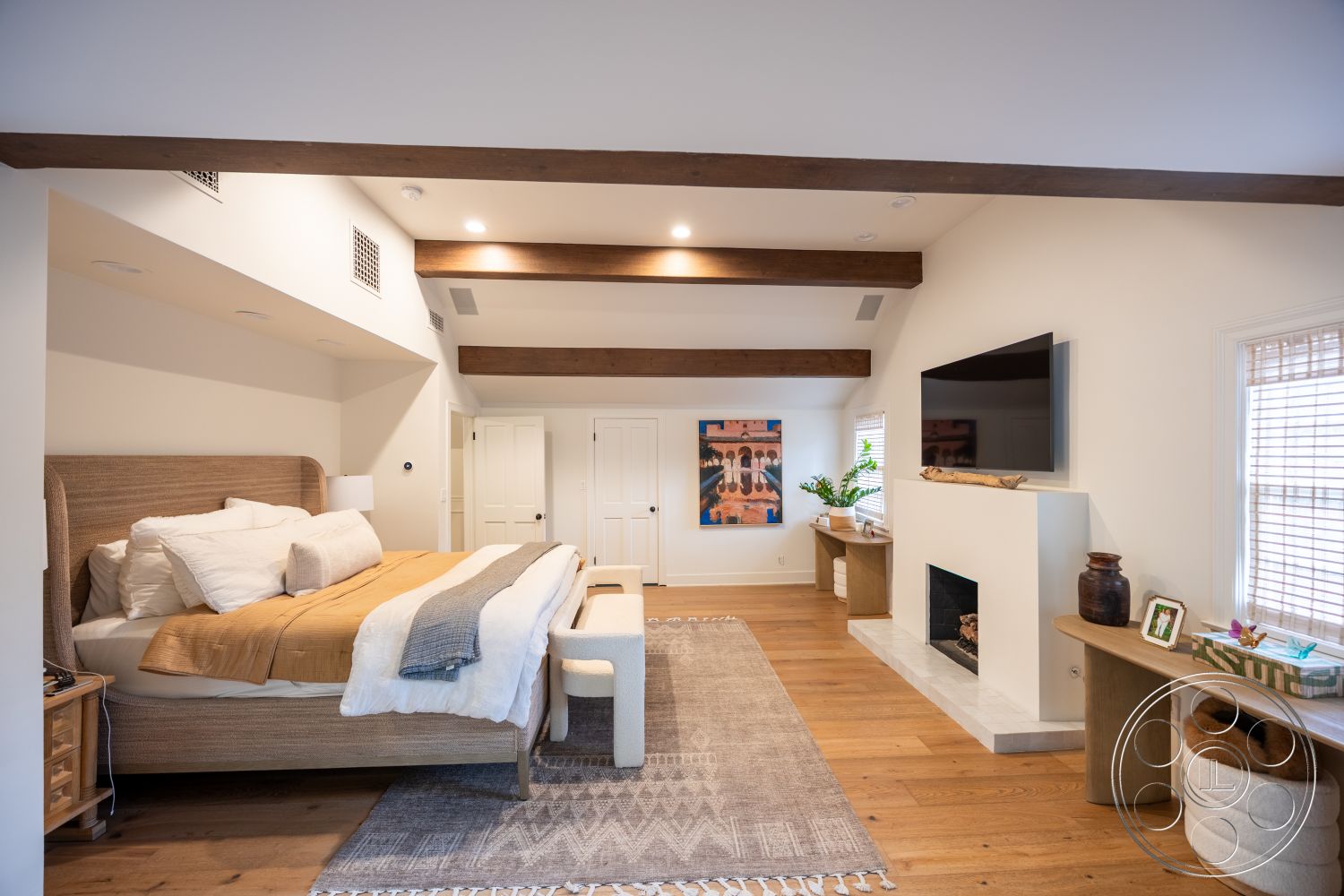 Americana 201 - neutral color palette, natural light from large windows, master bedroom, textured plaster walls, plush area rug, exposed wood ceiling beams, hardwood bedroom floors, minimalist decor style, built-in bedside cabinets, flat-screen television wall mount, modern fireplace with marble surround, stylish decorative accent pieces