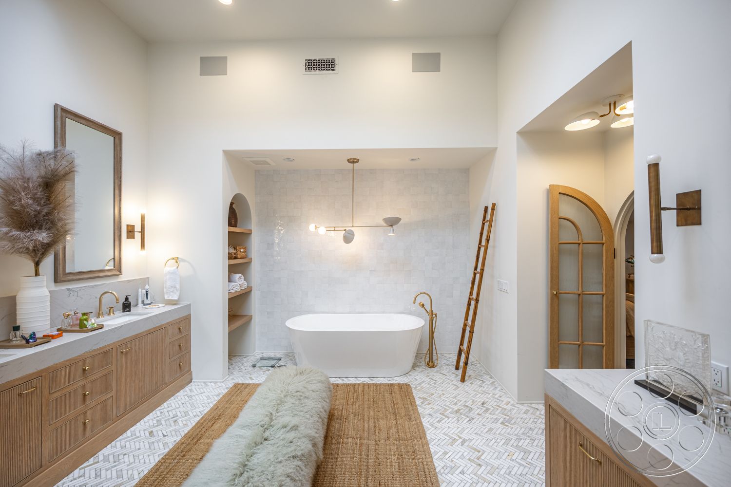 Americana 201 - neutral color palette, minimalist design elements, vaulted ceiling, arched doorway, modern light fixture, interior bathroom, freestanding soaking tub, ample natural light, marble tile bathroom walls, wooden vanity with gold fixtures, herringbone pattern tile flooring, textured woven area rug, backlit vanity mirror, glass shelving for toiletries, plush decorative items, decorative ladder for towels, ornate wall sconce lighting