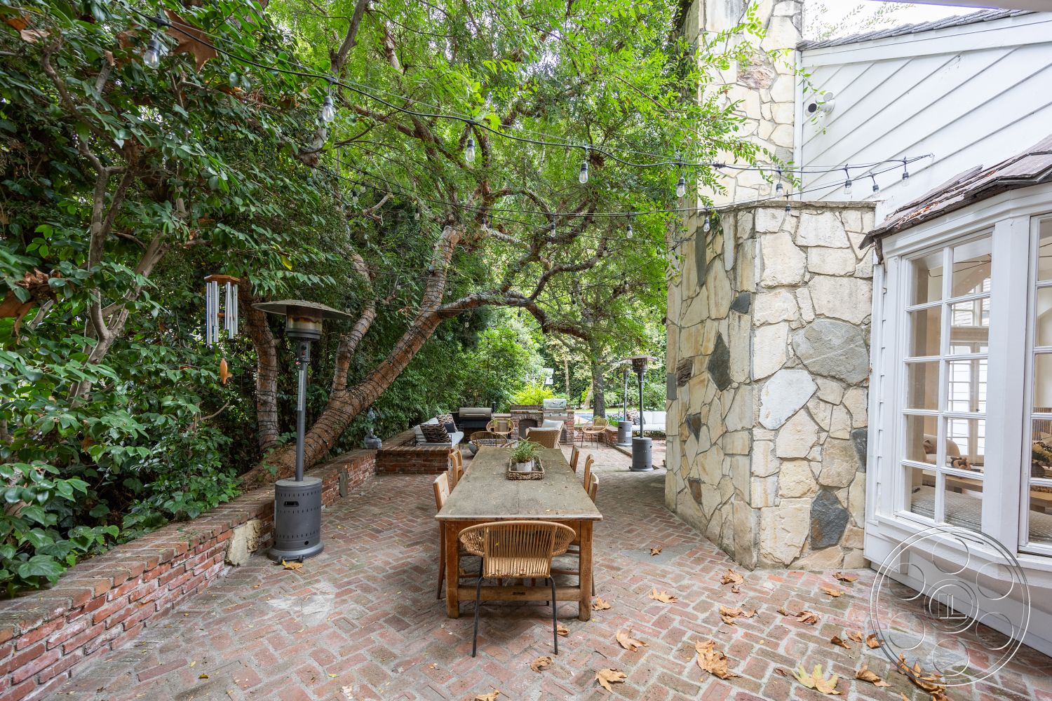 Americana 201 - exterior, stone accent wall, wooden dining table, wooden chairs, outdoor seating area, surrounding greenery, brick wall, natural stone pathway, tree canopies, metal patio heater, rustic outdoor decor, urban garden patio, brick patio flooring, relaxing garden setting, string lights overhead