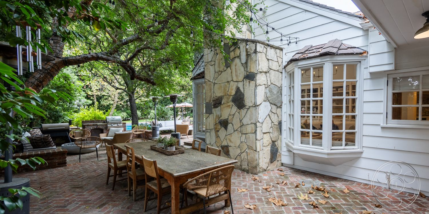 Americana 201 - exterior, wooden dining table, outdoor seating area, white clapboard siding, woven outdoor chairs, bay window, modern rustic design, stone feature wall, shingled roof, landscaped garden setting, brick patio flooring, overhanging tree branches, string lights for ambiance, adjacent pool area