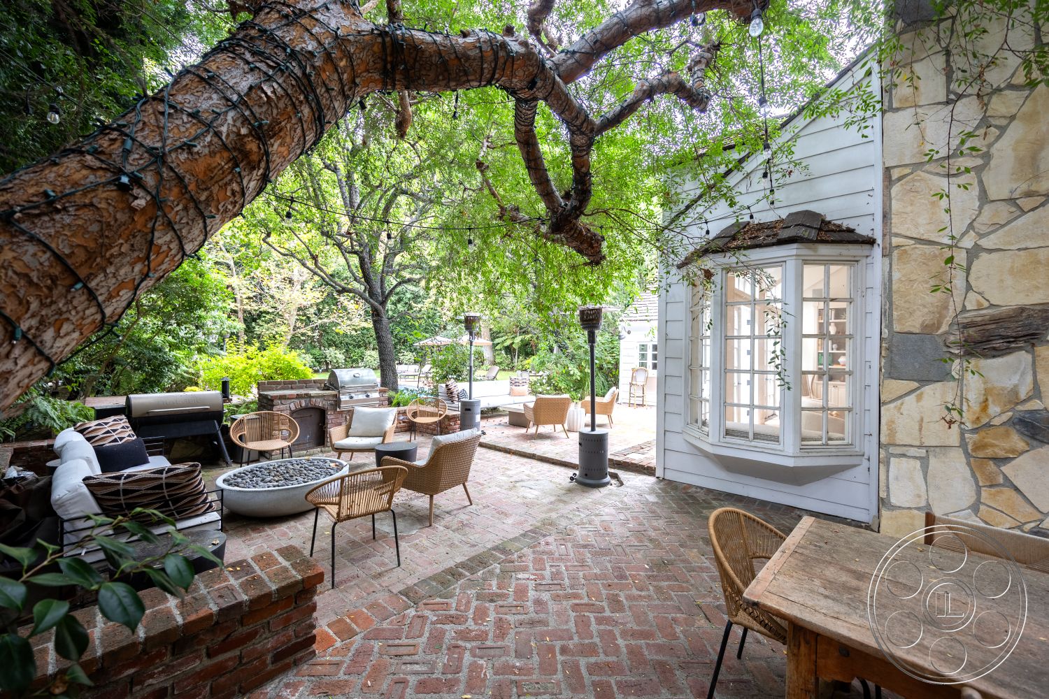 Americana 201 - modern outdoor furniture, landscaped backyard, outdoor fire pit, lush greenery surroundings, brick patio flooring, wooden outdoor dining table, brick barbecue area, interior window view, rustic outdoor cooking area, woven patio chairs, stone exterior wall, exposed brick retaining walls, green shaded garden setting, bay window with multiple panels, natural wood tree, decorative garden lighting, circular outdoor seating arrangement, urban garden escape