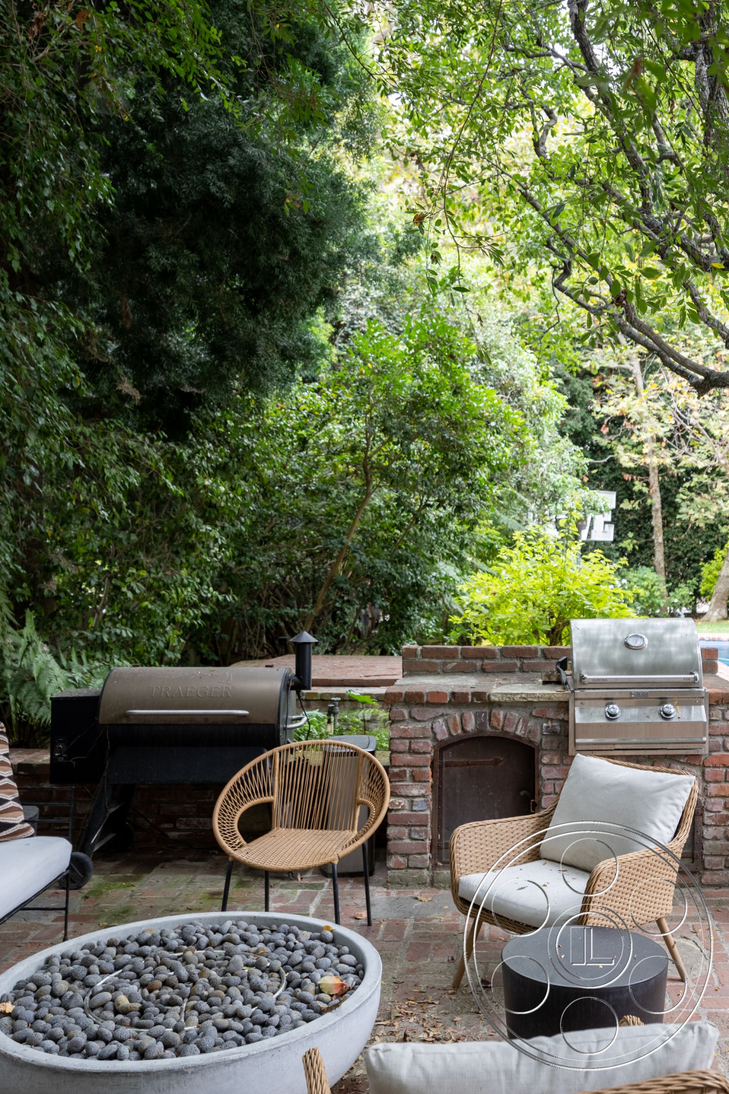 Americana 201 - surrounded by greenery, woven outdoor chairs, lush garden setting, brick patio flooring, modern round coffee table, brick outdoor barbecue area, circular fire pit with pebbles, exterior wooden accents, outdoor cooking space, rustic outdoor entertainment area