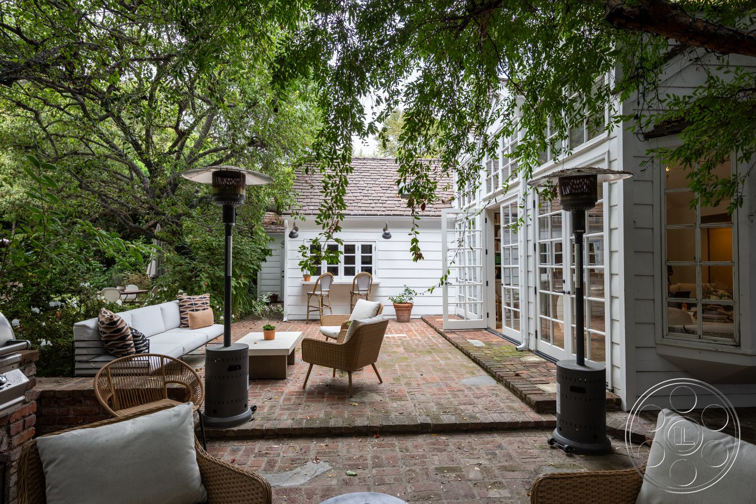 Americana 201 - exterior patio flooring with brick pavers, white wood panel siding, modern lounge furniture with fabric cushions, glass door access to interior living space, wooden pergola overhead with tropical plants, outdoor seating area with wicker lounge chairs, landscaped garden elements with lush greenery, outdoor dining space with table and chairs, patio heater installations for warmth, decorative planter with potted plants, quaint residential setting with leafy trees, vintage modern aesthetic style, open-air exterior common area, rustic charm with natural textures