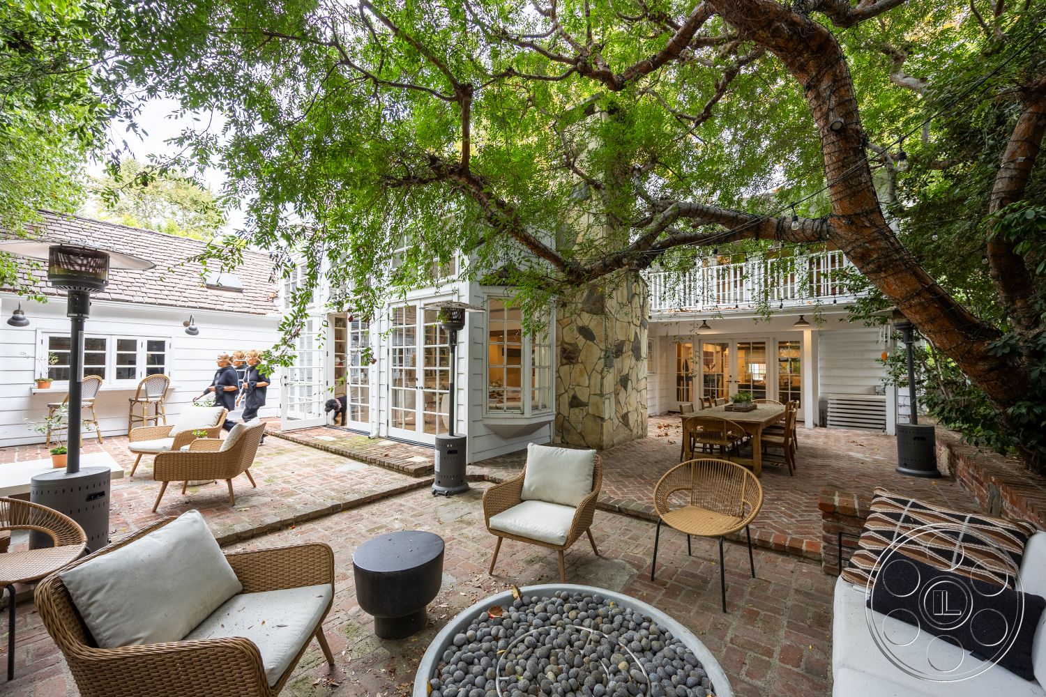 Americana 201 - exterior patio with brick flooring, exterior wooden deck railing, exterior natural stone feature wall, exterior white wood siding, exterior gabled roof with shingles, exterior built-in dining table, interior glass door openings, interior exposed wooden beams, interior large urban garden setting, interior cozy seating area with natural textiles, exterior landscaped with mature trees, interior warm ambient lighting, interior circular fire pit with pebbles