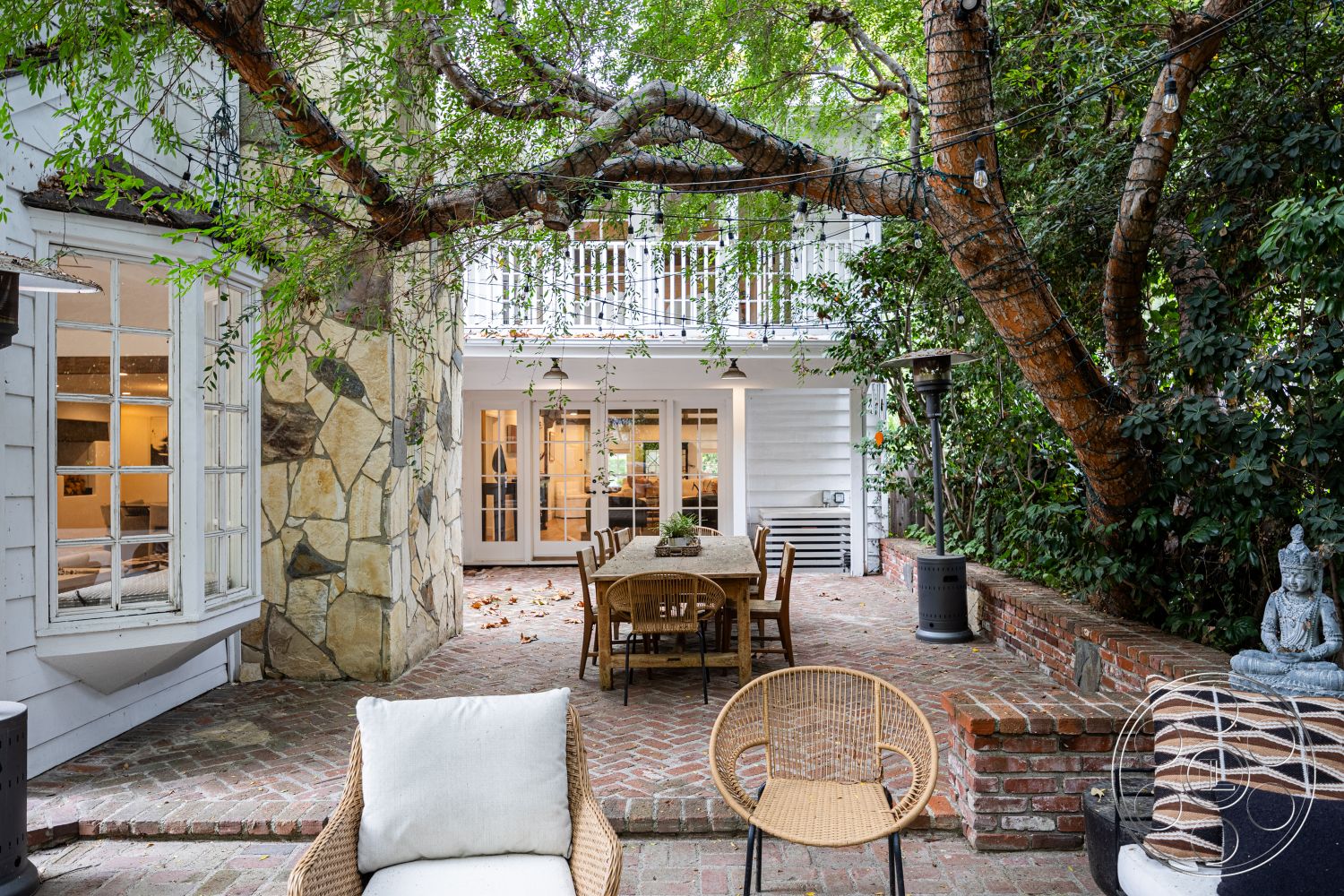 Americana 201 - lush greenery backdrop, modern outdoor furnishings, exterior brick patio flooring, exposed brick accent wall, decorative stone statue, suburban garden setting, natural stone wall cladding, white wooden window frames, shingle siding house exterior, wooden dining table on patio, outdoor seating area with rattan chairs, outdoor heating elements, string outdoor lighting, two-story residential architecture, traditional courtyard design