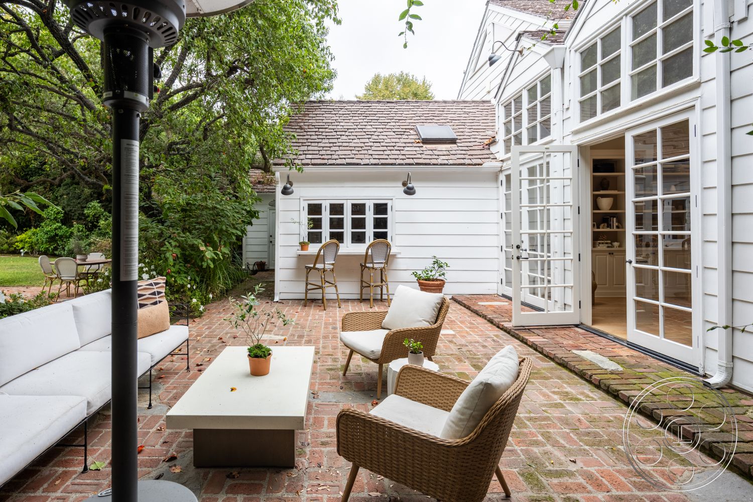 Americana 201 - wooden dining chairs, open-air layout, concrete coffee table, landscaped garden setting, exterior brick patio flooring, French doors with glass panels, outdoor seating area with wicker chairs, white wooden siding walls, sloped roof with shingles, lush greenery surrounding the patio, modern outdoor heater, single-story home with additional wing, stylish outdoor aesthetic, rustic charm ambiance