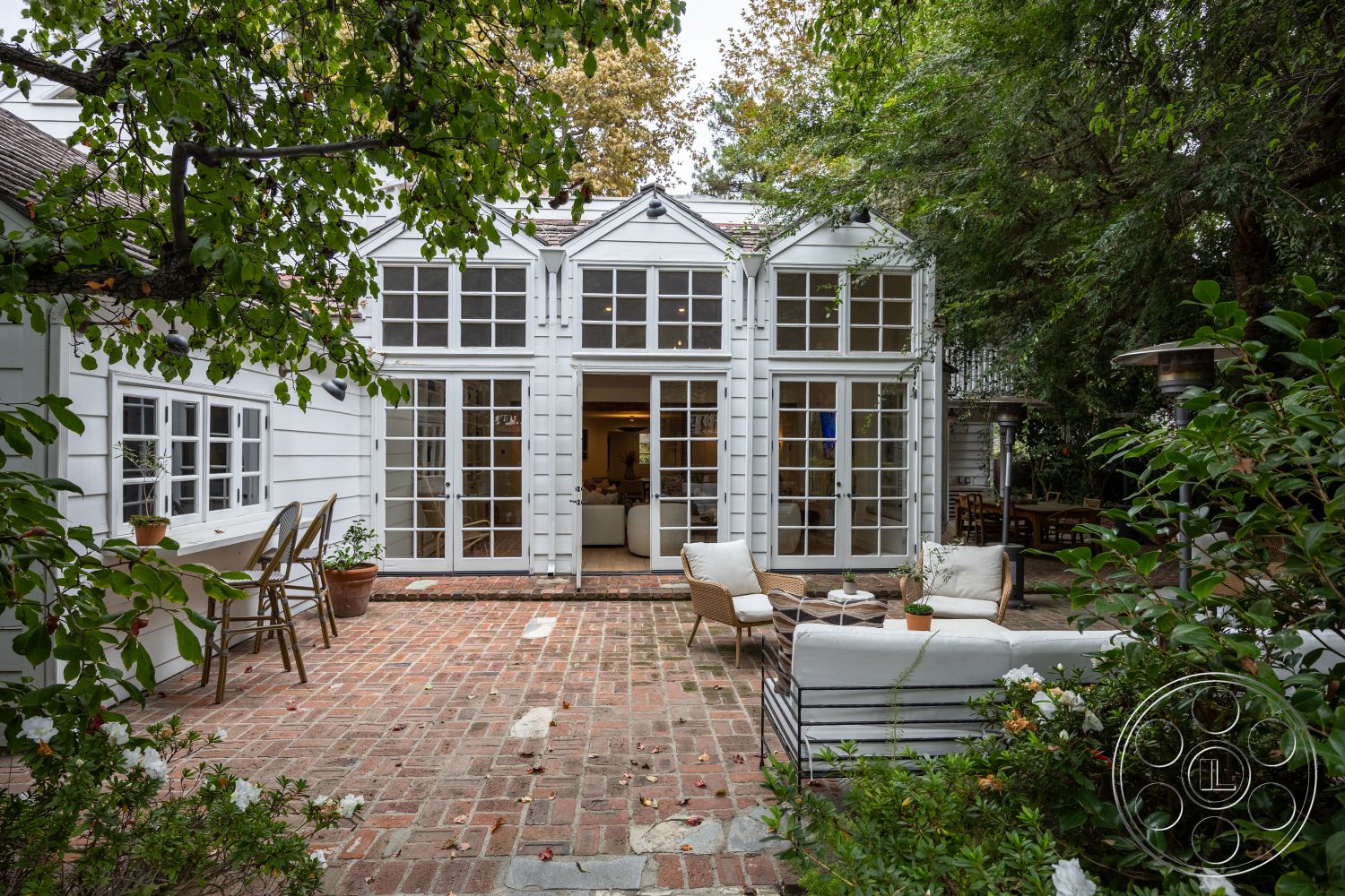 Americana 201 - exterior, outdoor dining area, garden setting, modern rustic style, double French doors, natural stone path, arched windows, brick patio flooring, rustic outdoor seating, white wood panel siding, overhead gabled roof, green foliage landscaping, textured brick patio surface, inviting courtyard, wood railing accents