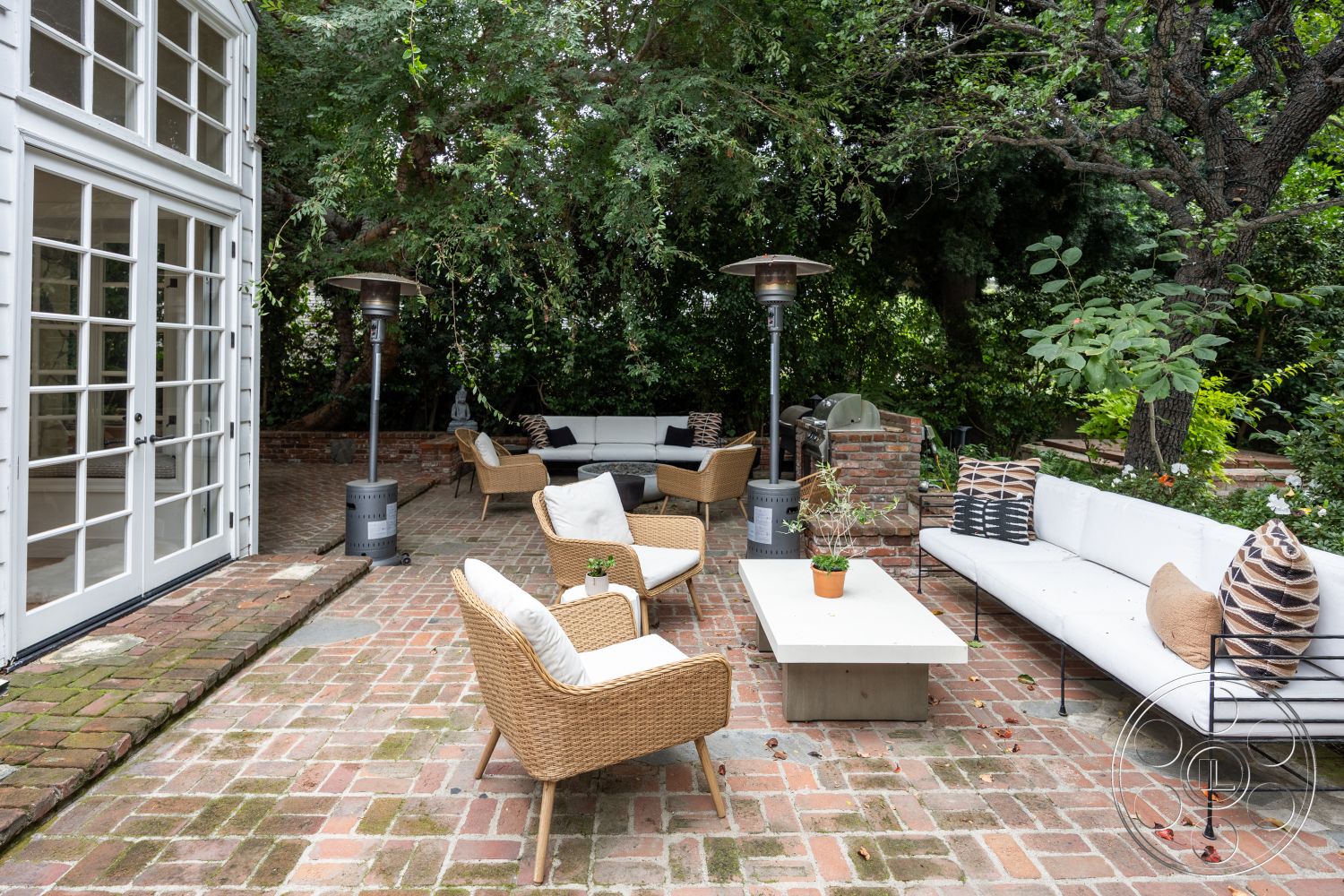 Americana 201 - textured throw pillows, decorative outdoor lighting, concrete coffee table, lush garden setting, modern outdoor seating area, exterior patio with brick flooring, cozy outdoor living space, open-air gathering space, glass French doors, woven rattan lounge chairs, domestic urban backyard, stone and brick grill area, greenery landscape with trees, seating arrangement under foliage