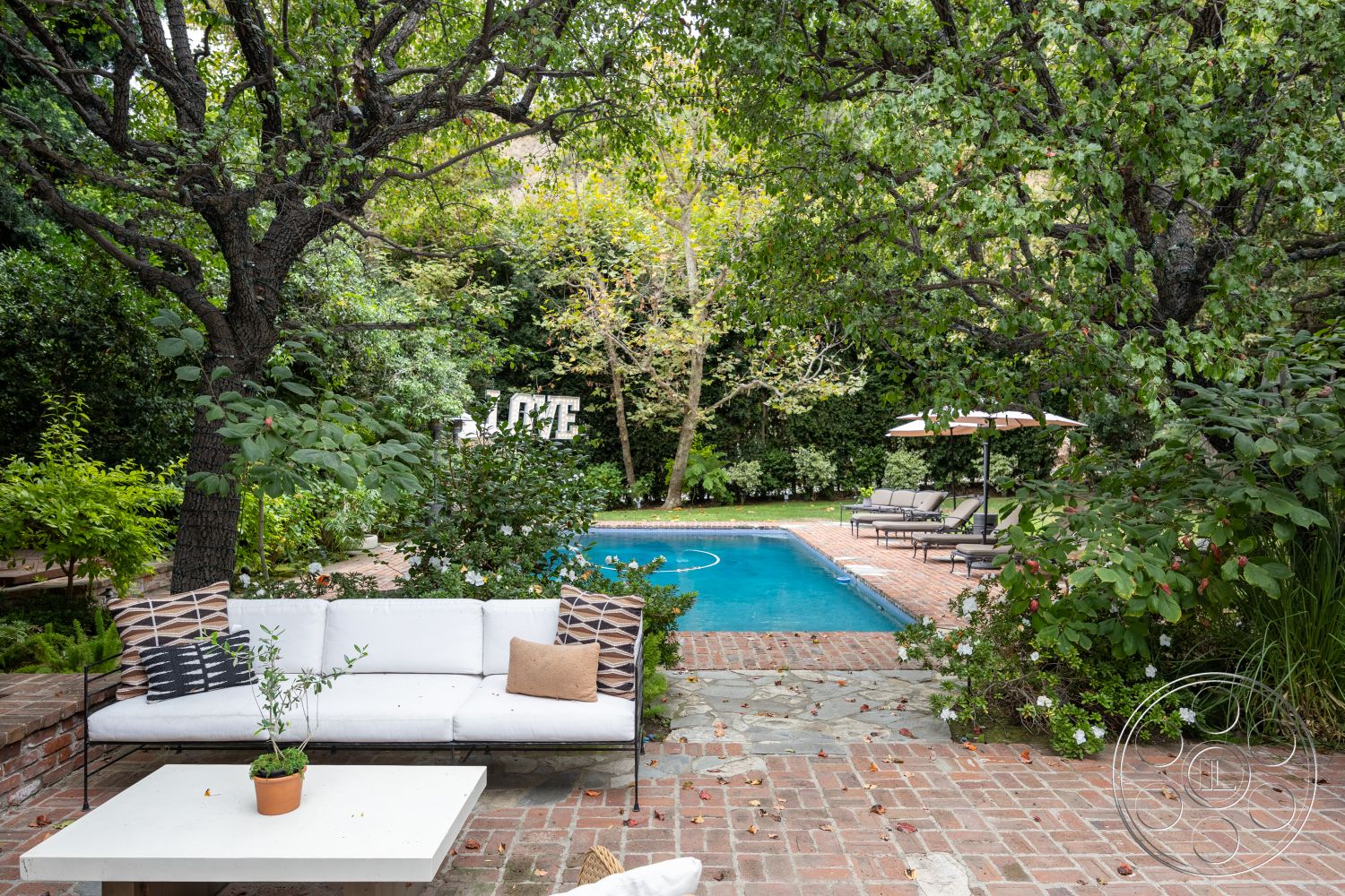 Americana 201 - landscaped yard, rectangular swimming pool, brick pathway, outdoor lounge area, metal patio furniture, surrounded by lush greenery, brick patio flooring, natural stone pool deck, exterior pool area, surrounding trees and foliage, decorative outdoor cushions, tropical garden setting, wooden table with potted plants, shaded by trees, sun loungers with umbrellas, hardwood lounge couch