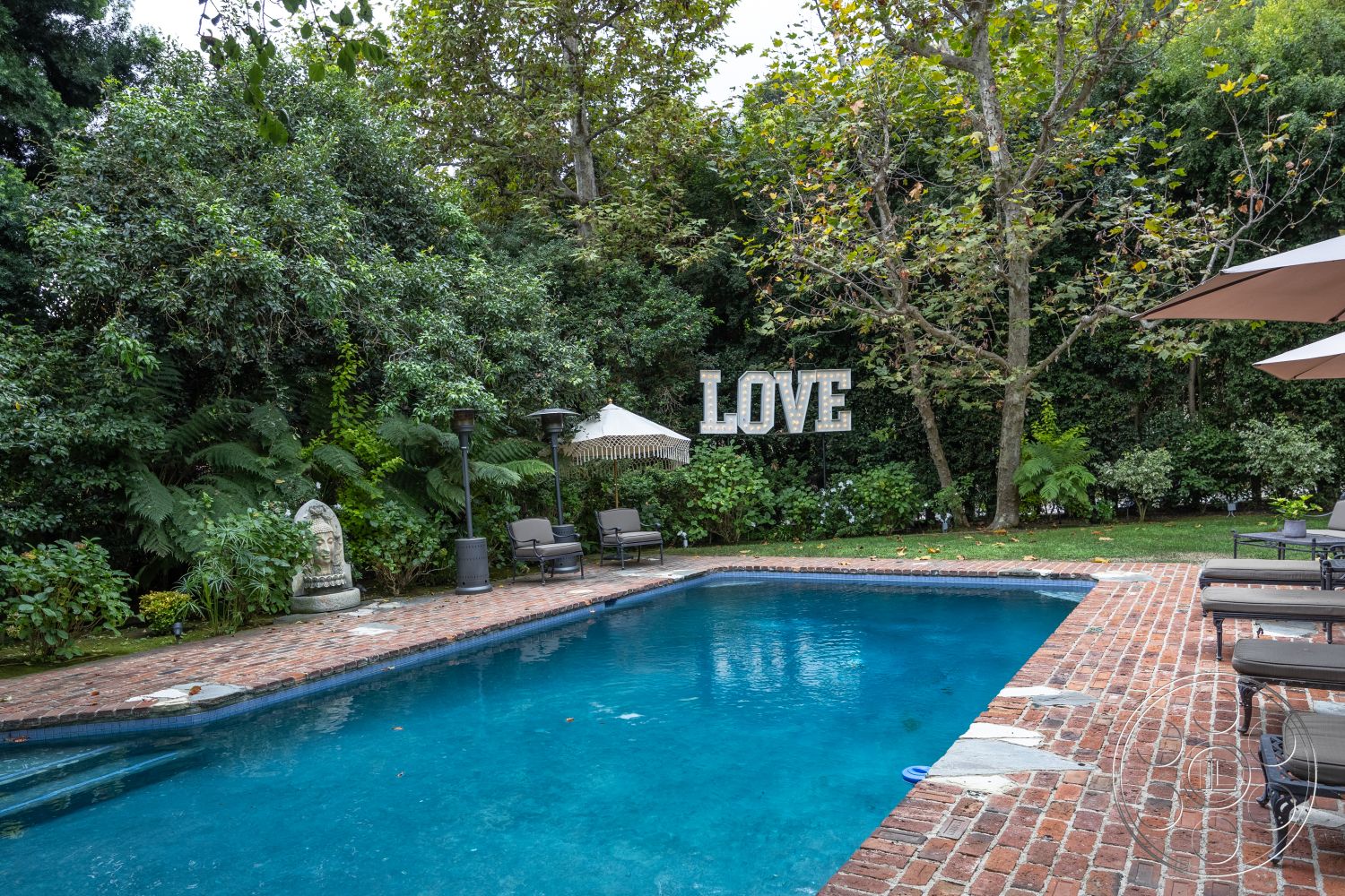 Americana 201 - rectangular swimming pool, lush greenery backdrop, landscaped garden setting, concrete pool coping, exterior brick pool deck, outdoor patio lounge area, wooden patio umbrella, outdoor space with seating, stone decorative statue, decorative 'LOVE' sign, brick paver pathway, patio area with lounge chairs, surrounding trees for shade, sunny outdoor relaxation space