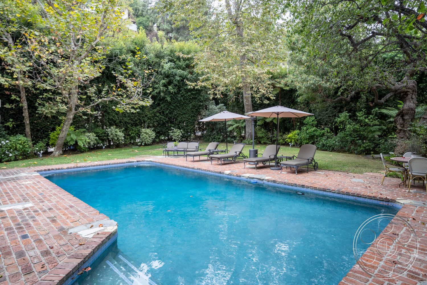 Americana 201 - surrounding greenery, brick pool deck, rectangle swimming pool, landscaped backyard with hedges, patio area with lounge chairs, inground pool with concrete border, sunshade umbrellas on patio, natural stone accents around pool, outdoor garden seating, exterior setting with trees